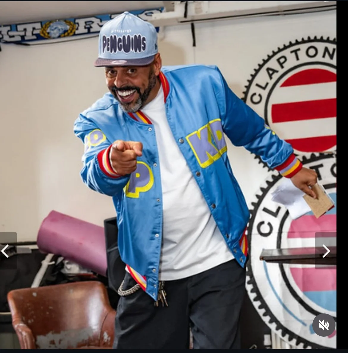 A smiling man wearing a blue varsity jacket with yellow Greek letters, a gray cap with the word 'Pelicans,' and a chain, pointing toward the camera. Behind him is a wall with the Claxtony logo.