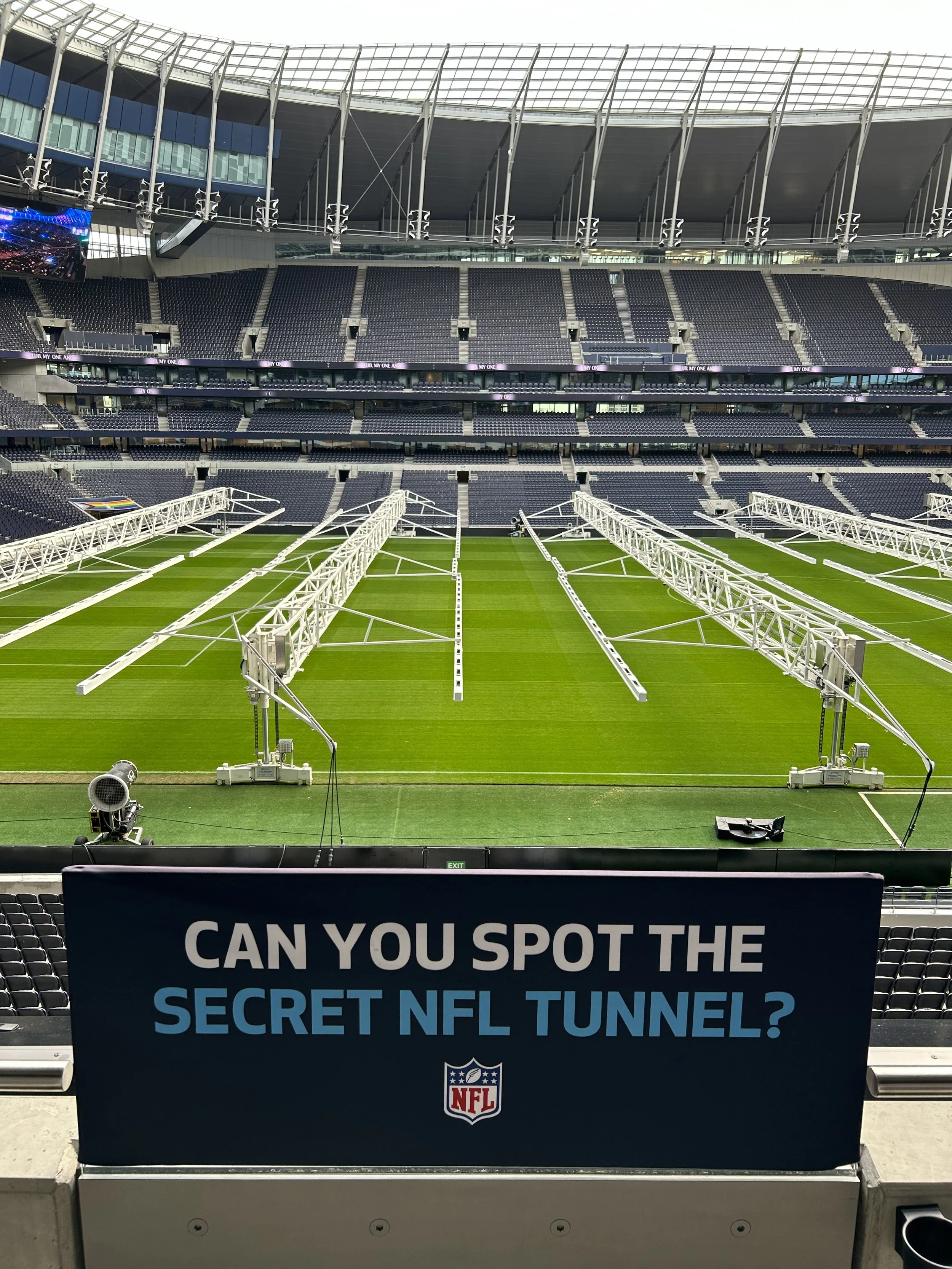 View of a football stadium with empty seats, lighting structures, and a partially assembled NFL tunnel on the field, with a sign in the foreground asking if viewers can spot the secret NFL tunnel.
