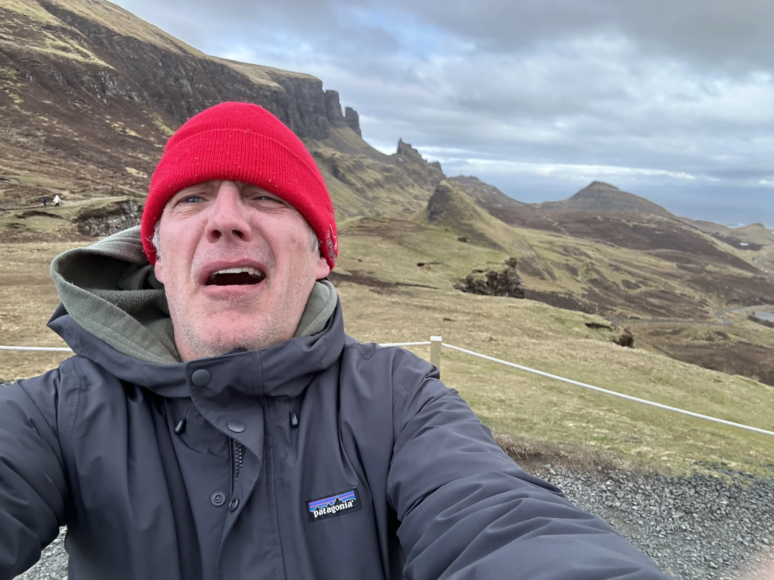 Man taking a selfie outdoors with a mountainous landscape in the background, wearing a red beanie and a Patagonia jacket.