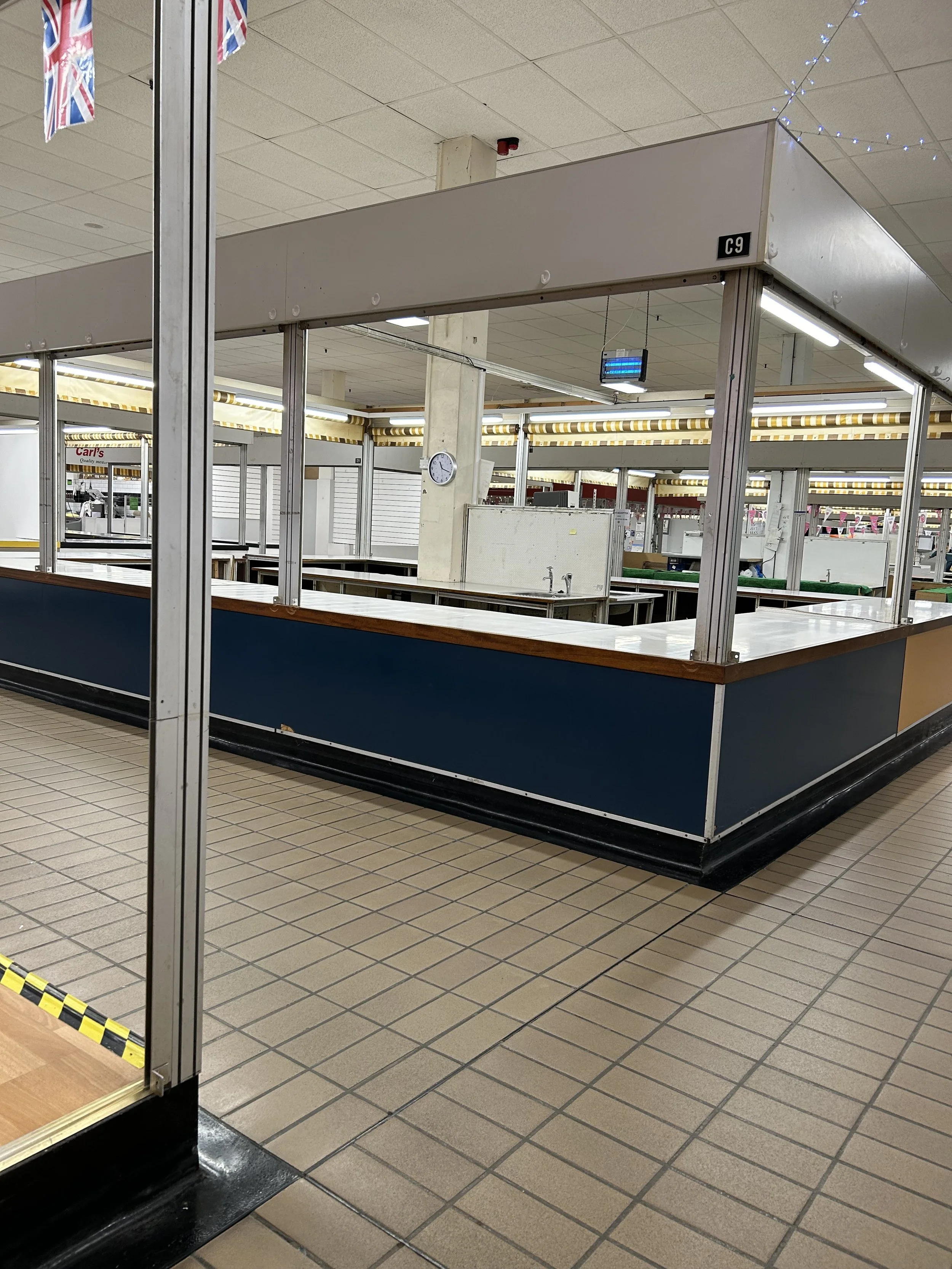 Empty market stall with a blue lower panel, metal support structure, and wooden countertop, inside a store with tiled floors and ceiling, fluorescent lighting, and UK flags hanging above.