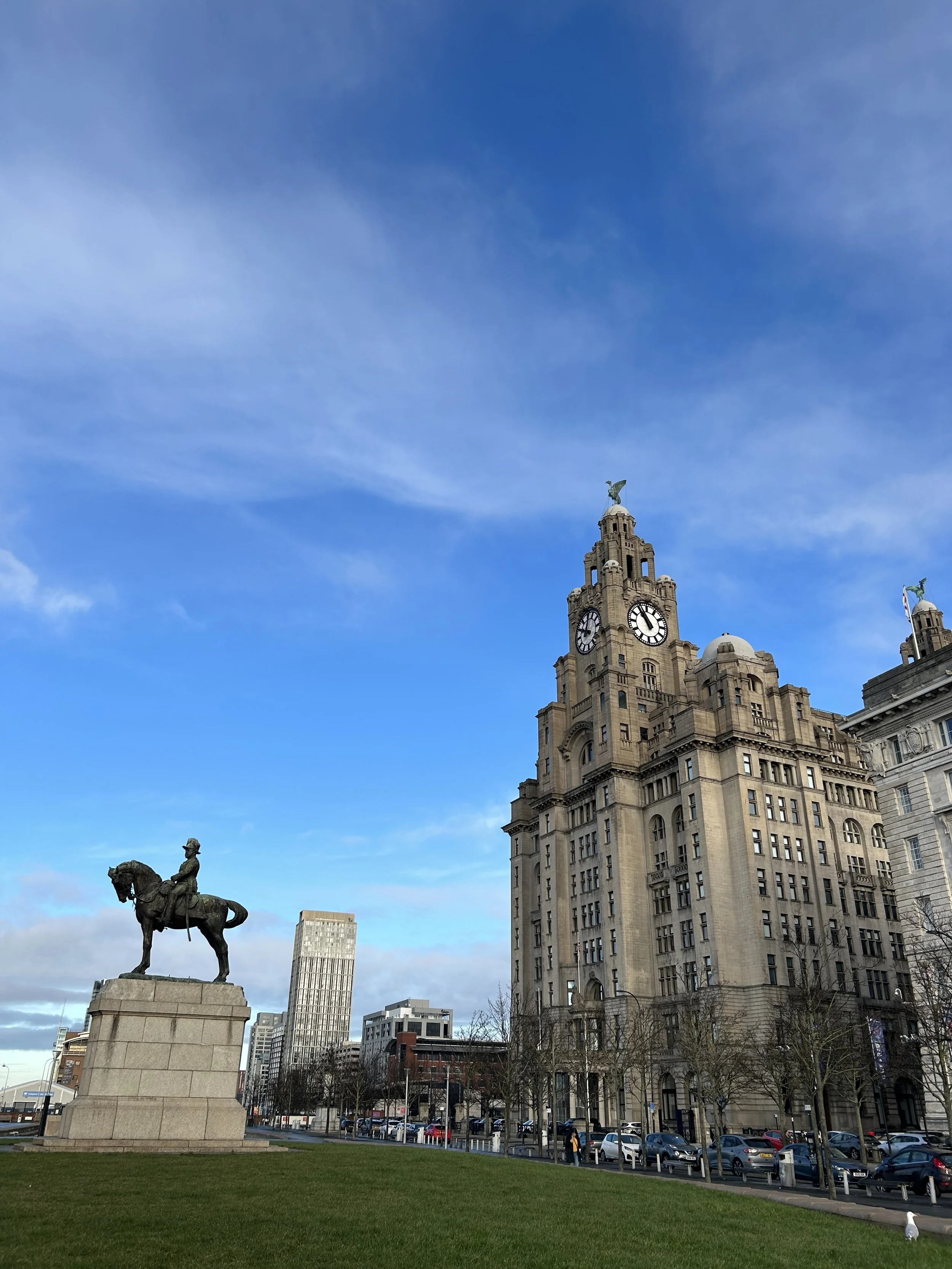 A historic clock tower building with a statue of an eagle on top, next to an equestrian statue in a park with trees, cars, and buildings in the background under a blue sky with clouds.