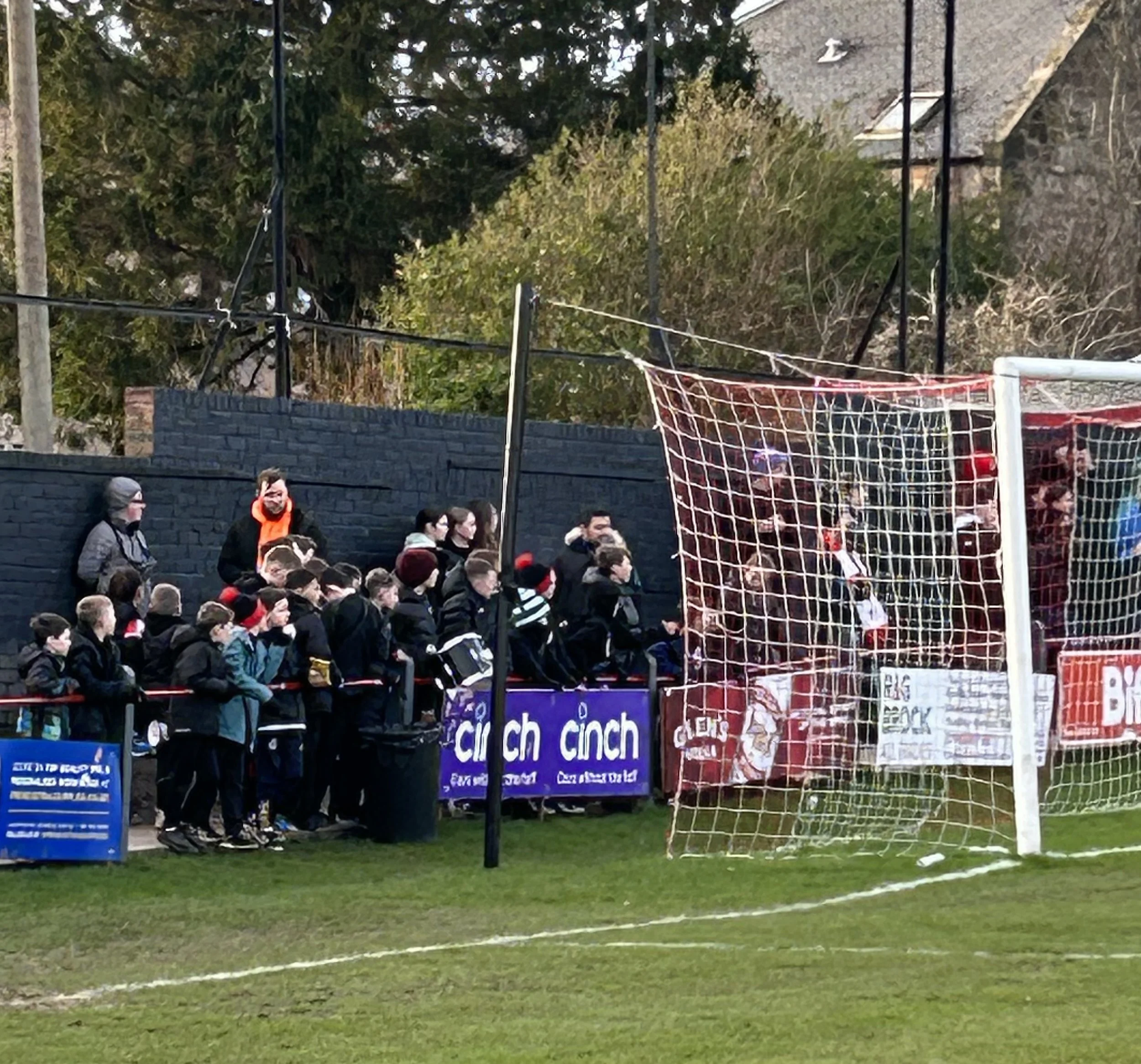 Children and adults watching a youth soccer game from behind the goal net on a field, with advertising banners on the wall behind them and trees in the background.