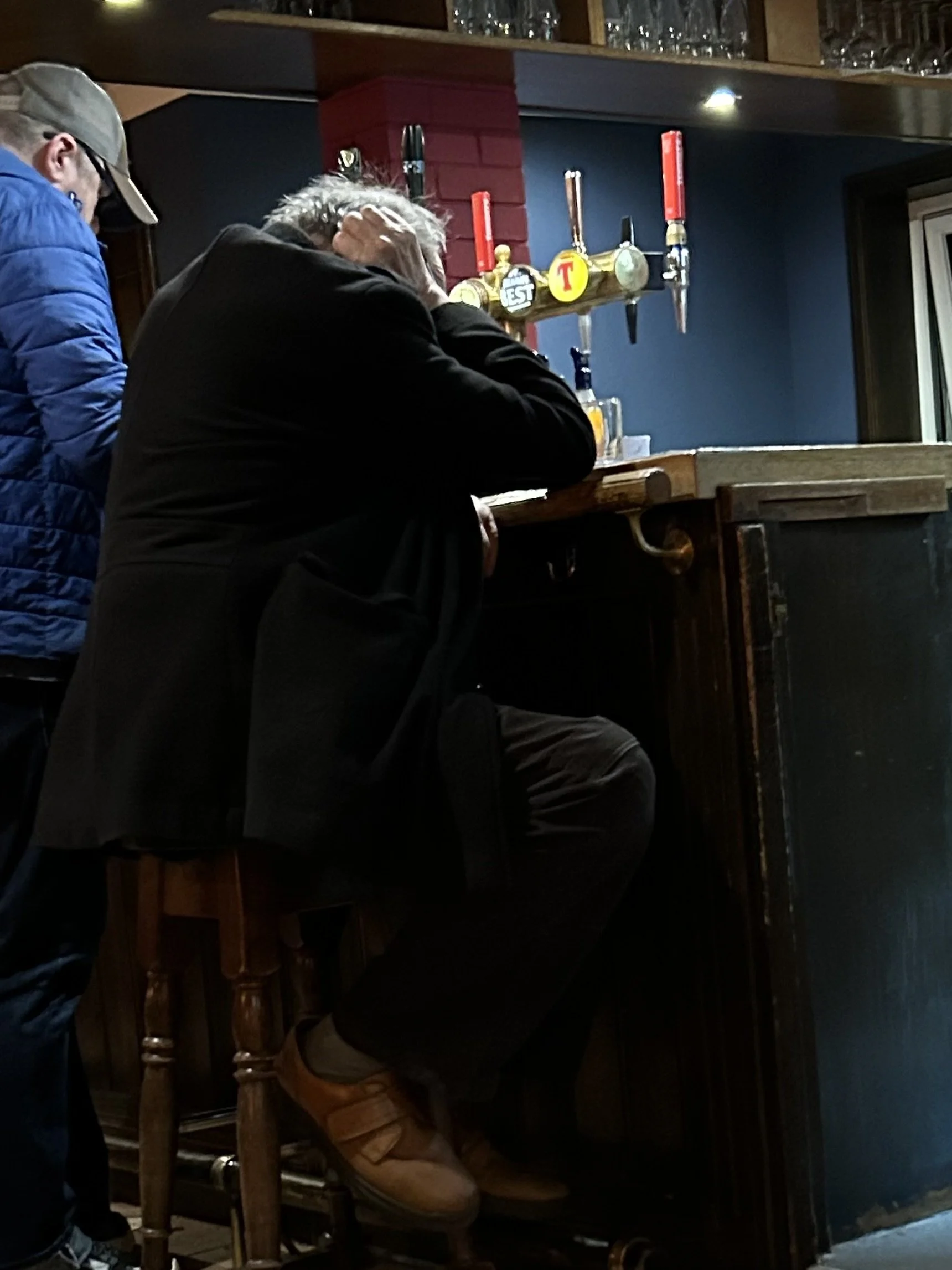 An older man with gray hair and a black jacket is sitting at a bar, resting his head on his hand. A younger man in a blue jacket and baseball cap is standing nearby. The bar has a red brick wall and beer taps.