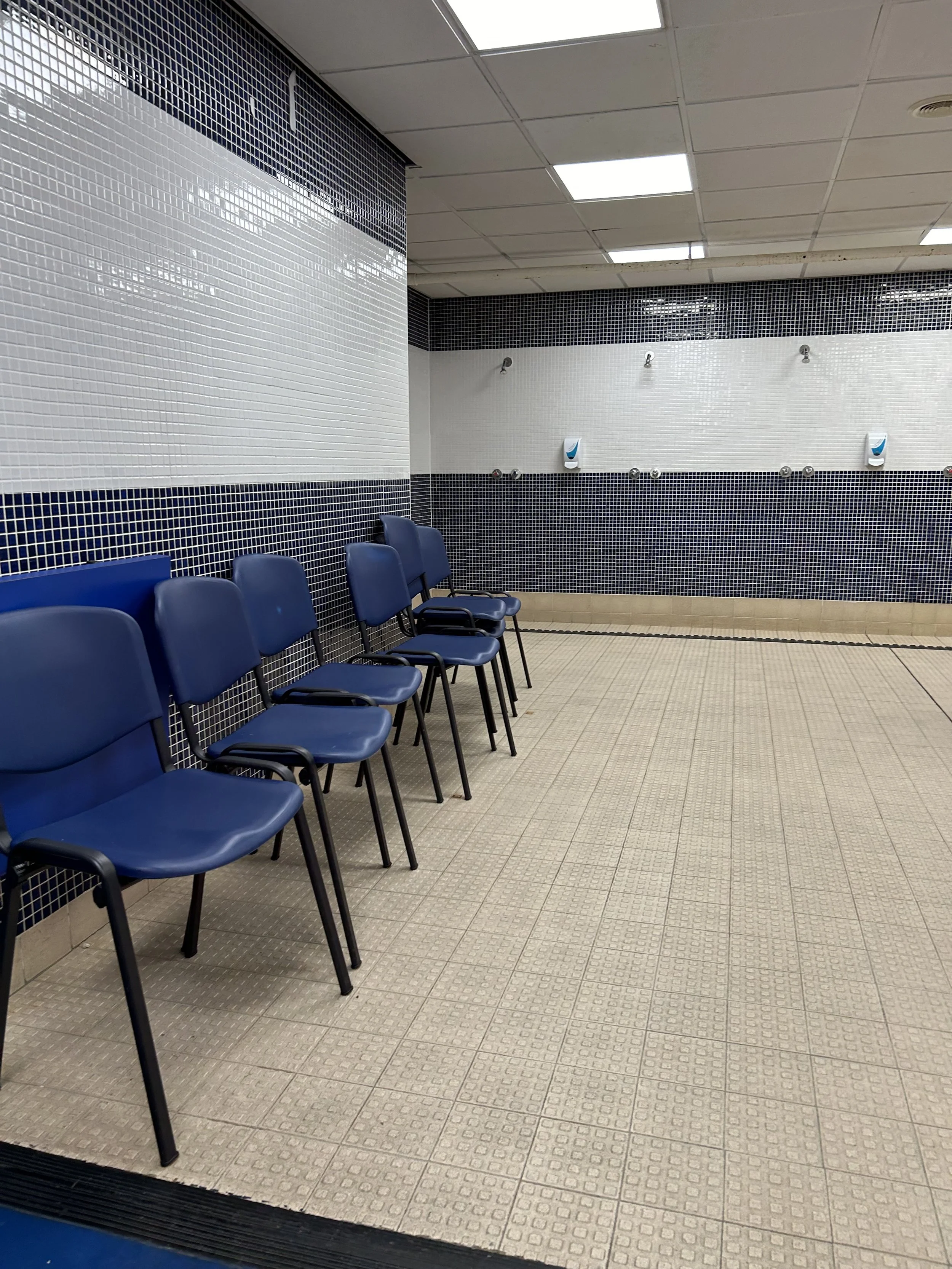 Empty waiting area with six blue chairs along a tiled wall in a public facility, possibly a medical or government office, with tiled walls and ceiling lights.