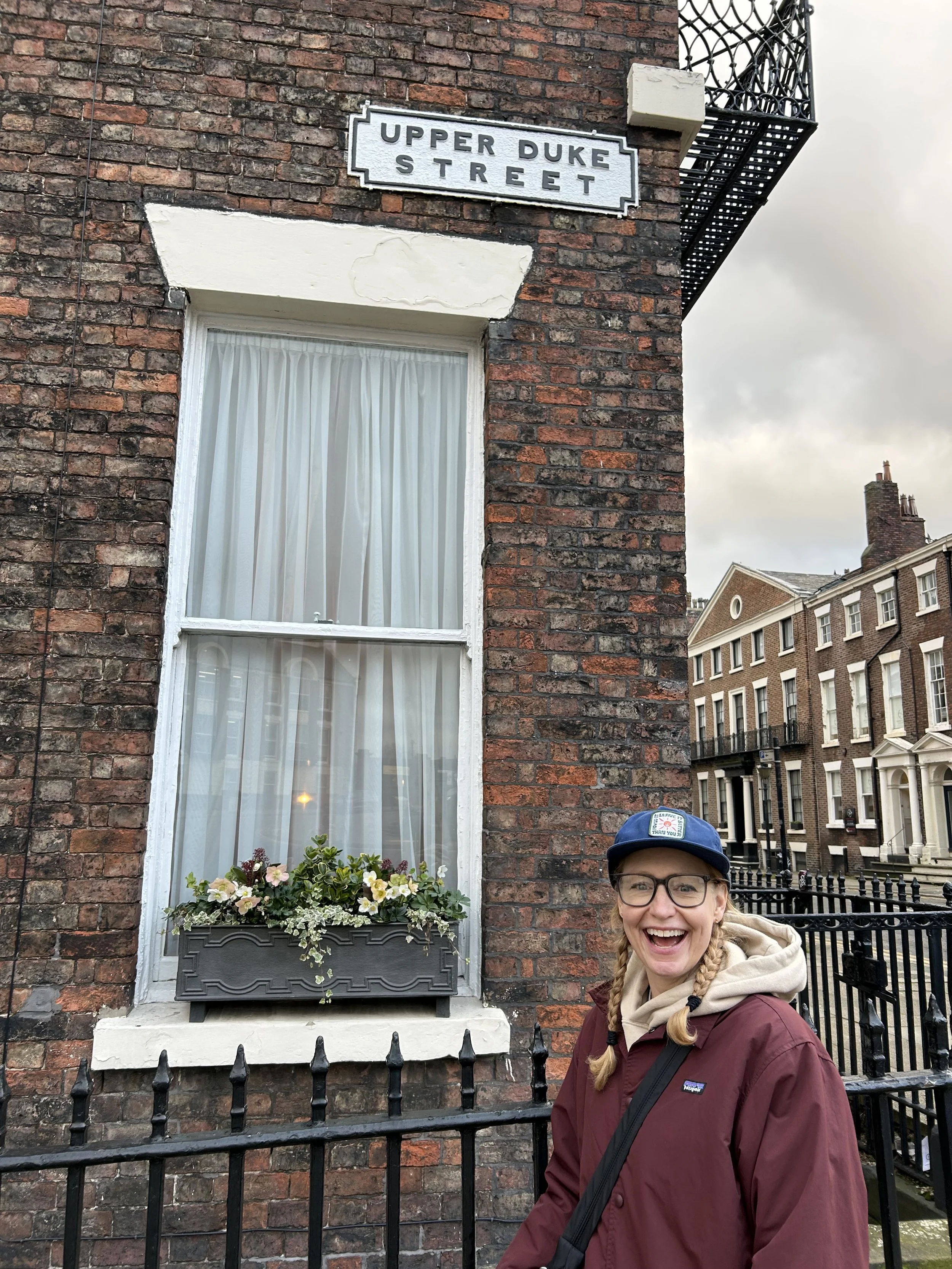 A smiling woman with glasses, wearing a blue cap, a maroon jacket, and a beige hoodie, standing outside in front of a brick building with a window adorned with a flower box. The building has a street sign that reads "Upper Duke Street."