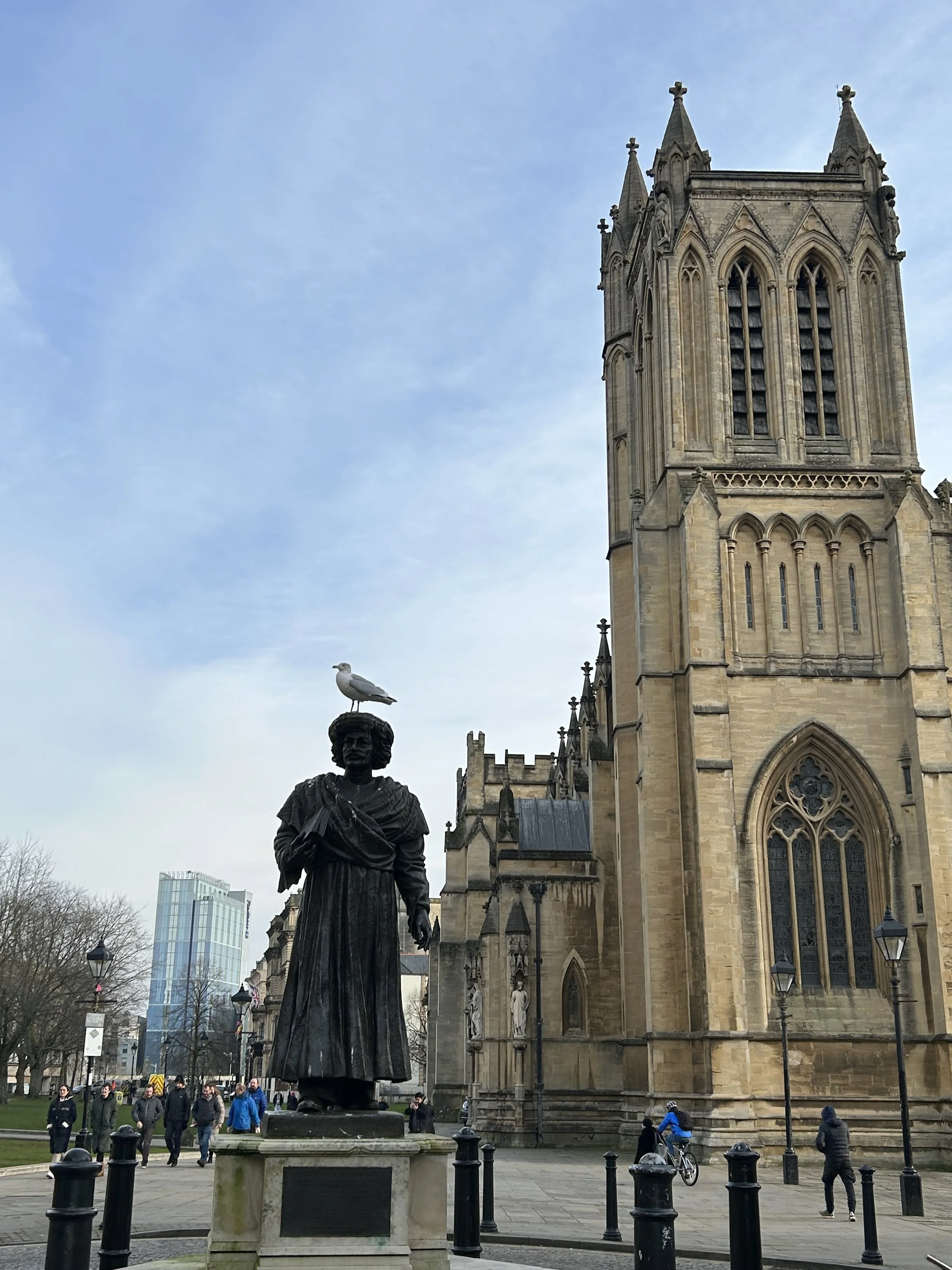 A statue of a woman with a bird perched on her head in front of a historic cathedral with tall towers and stained glass windows, surrounded by people and street lamps.