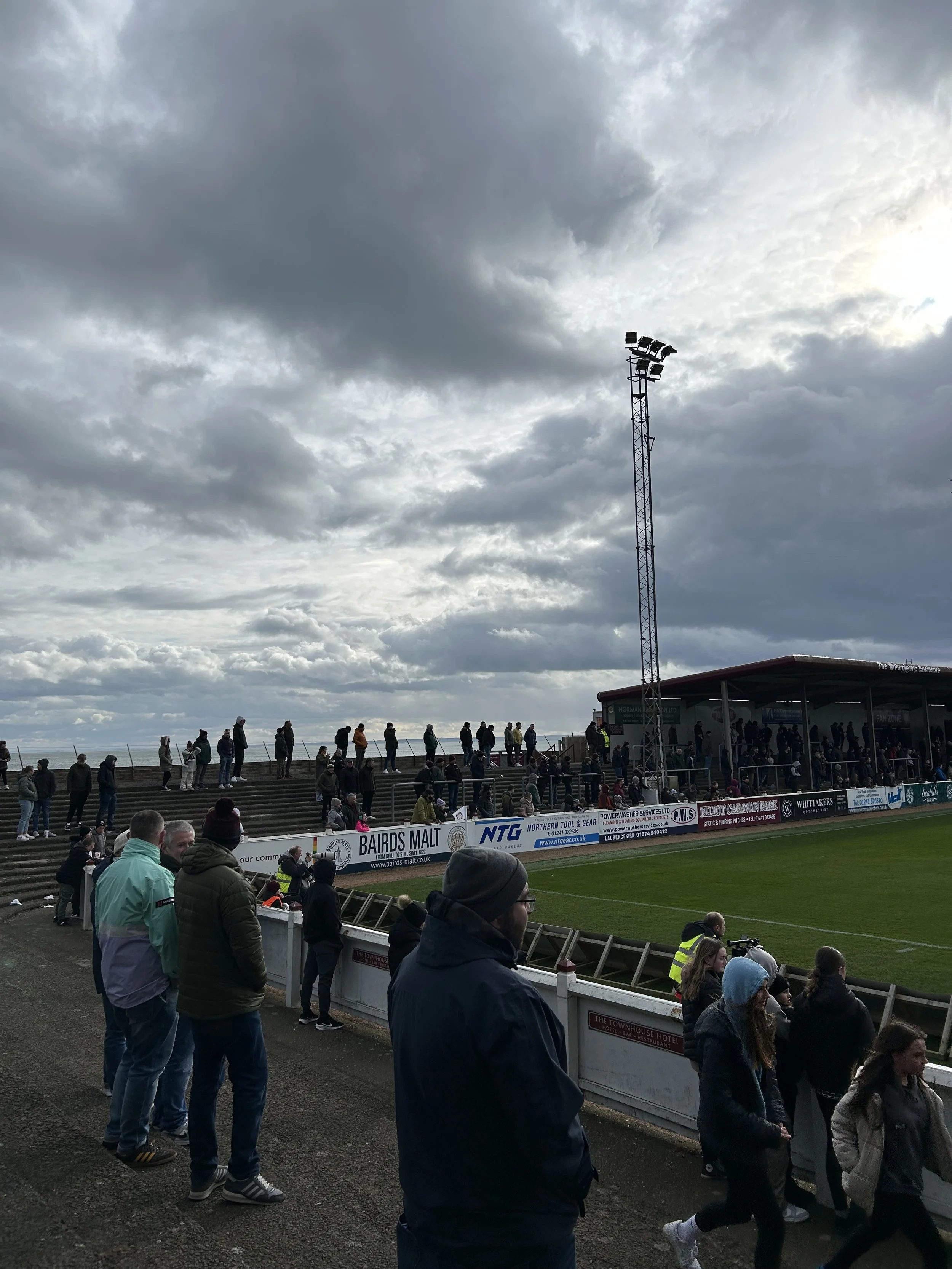 People watching a sporting event at a stadium on a cloudy day, with some spectators seated on steps and others standing near the edge of the field.