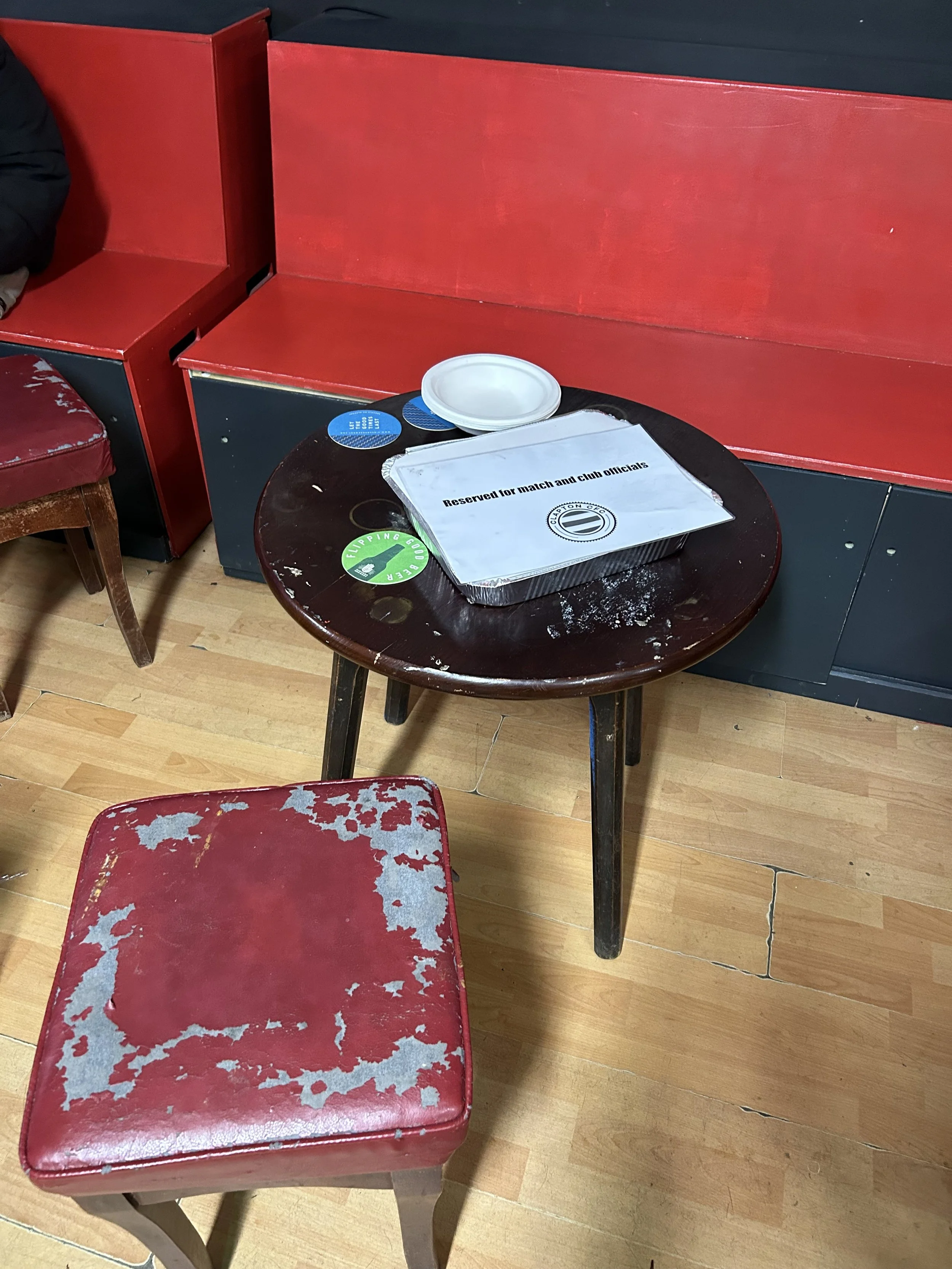 A round wooden table with a reserved sign, a small disposable food container, a white paper plate, and a couple of blue coasters. Surrounding the table are red and black cushioned benches and chairs with worn, peeling red upholstery on the seats, sit