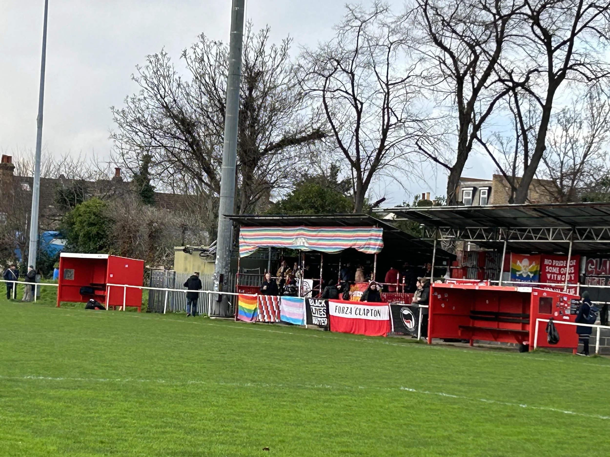 A soccer field with a raised spectator area, banners, and flags supporting Black Lives Matter and LGBTQ rights, with leafless trees and houses in the background.