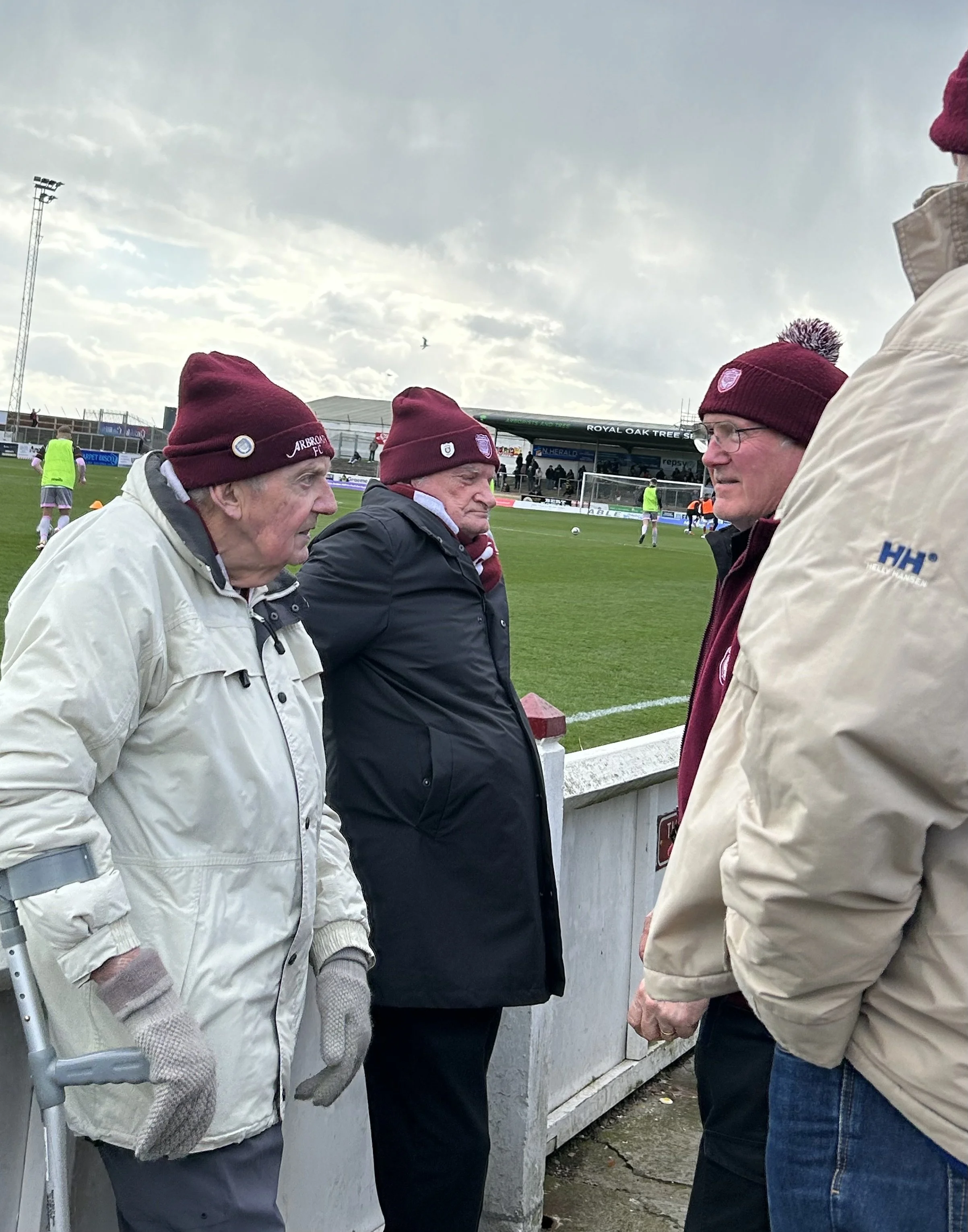 Four elderly men wearing maroon beanies and jackets, standing at a football field's sideline, engaged in conversation during a cloudy day.
