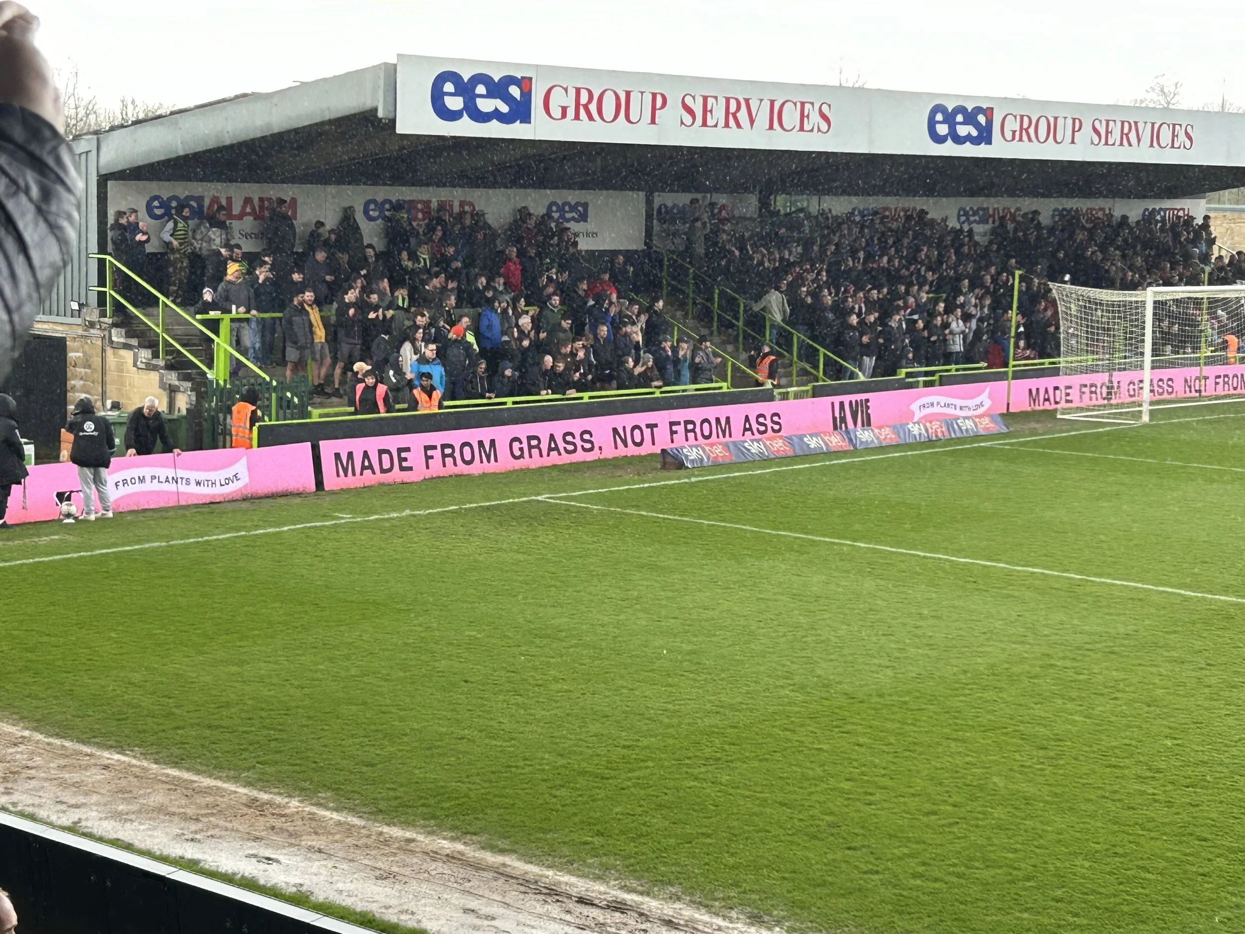 A soccer stadium with spectators seated in a covered stands area. The field is green with white lines. There is an electronic advertising banner along the sideline displaying the message 'MADE FROM GRASS, NOT FROM ASS' in pink background.