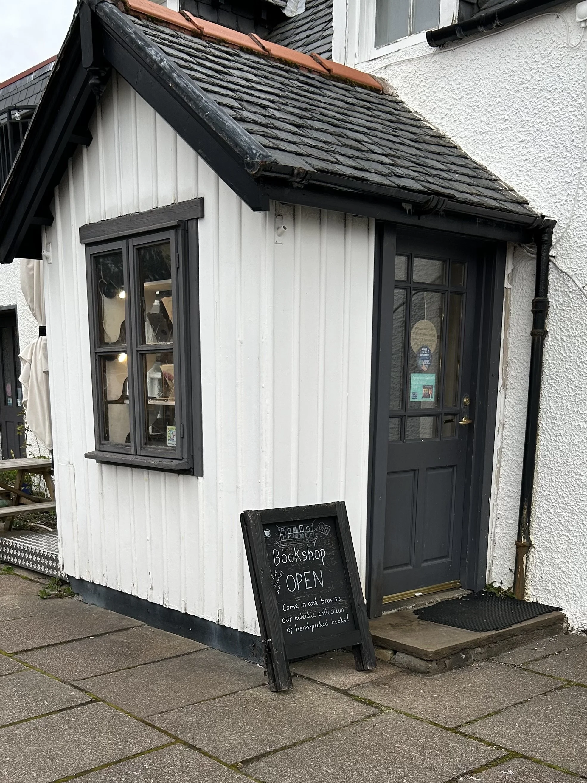 Small white wooden building with black window frames and a black door, serving as a bookstore. A chalkboard sign outside reads 'Bookshop OPEN' and invites customers to browse a collection of hand-packed books.