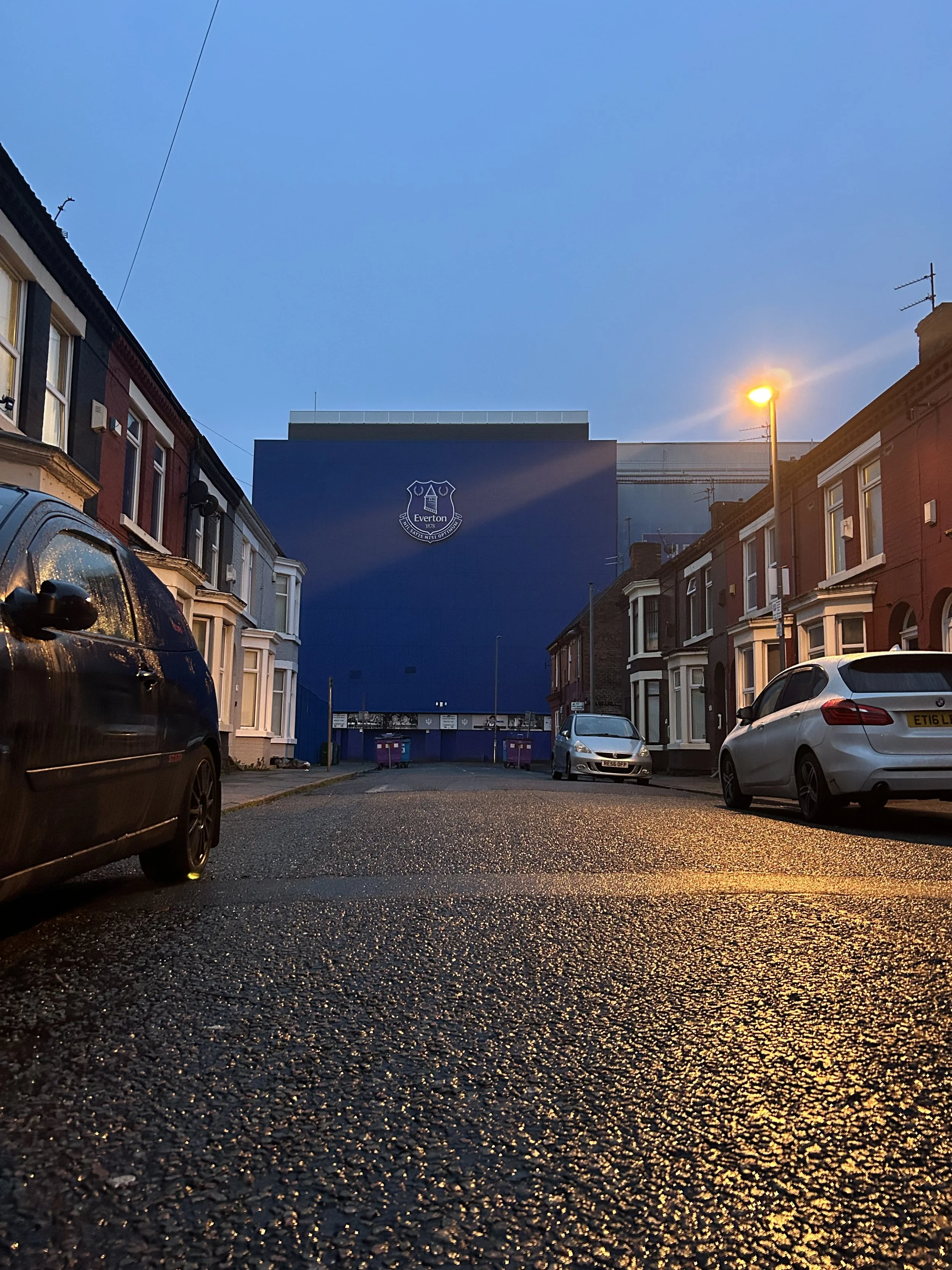 A residential street during evening with parked cars, typical row houses, and an Everton football club building visible in the background.