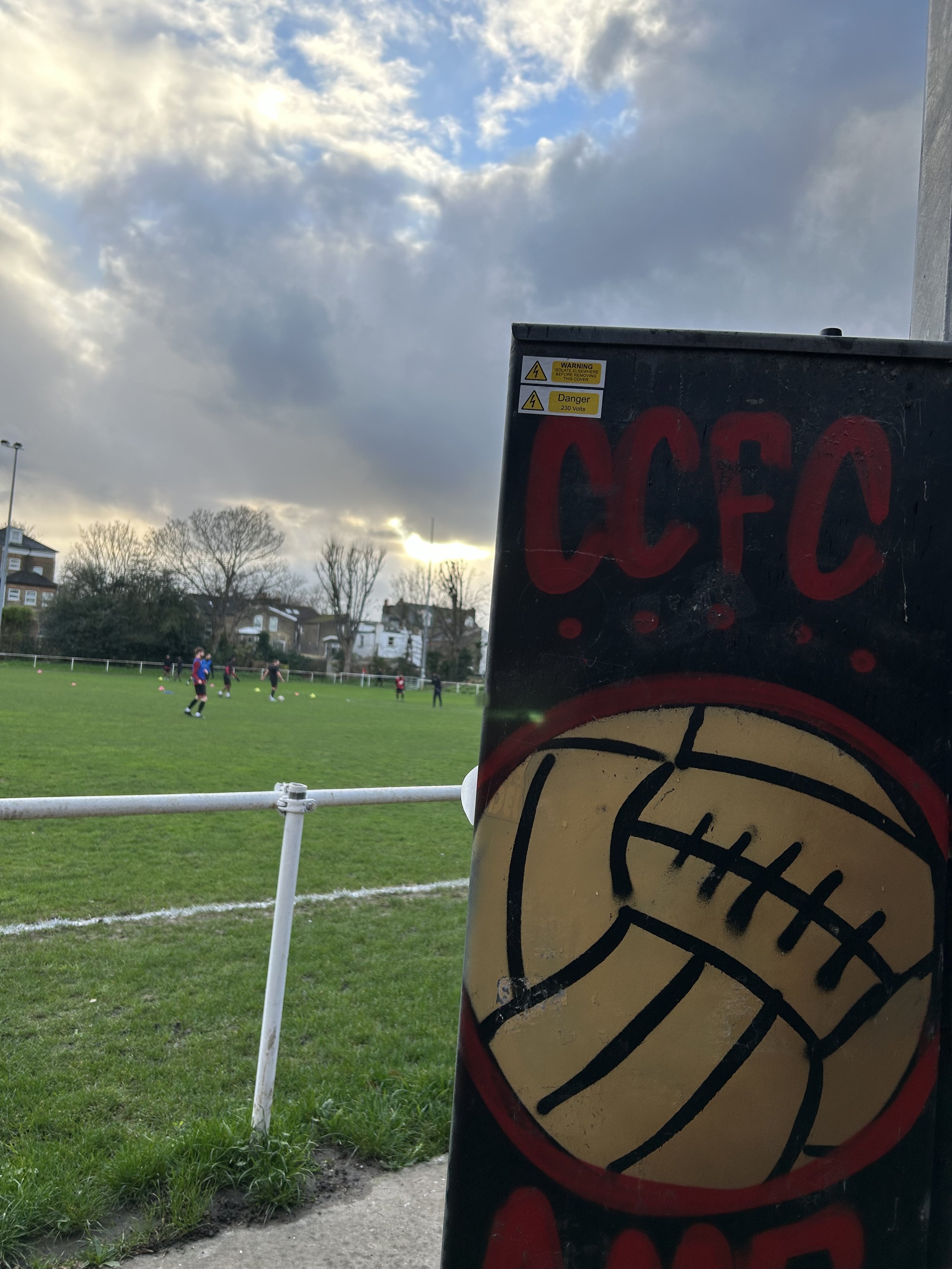 A grassy sports field with soccer players and a line of trees in the background, viewed through a metal fence and partially obscured by a black utility box featuring red graffiti and a yellow sticker with a black football illustration.