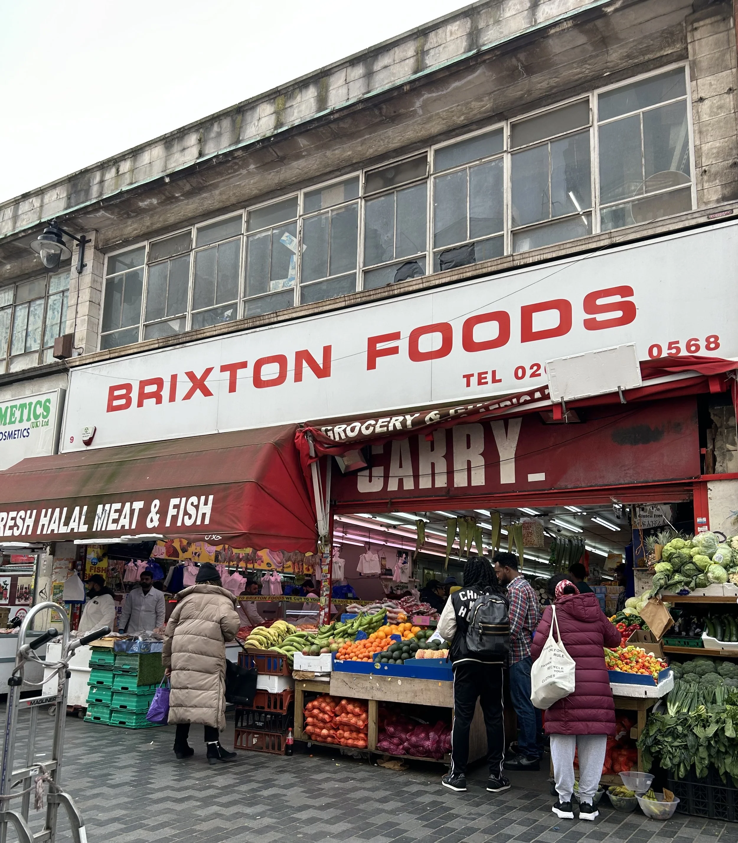 A lively street market scene outside Brixton Foods with shoppers buying fresh fruits and vegetables, under a red awning.