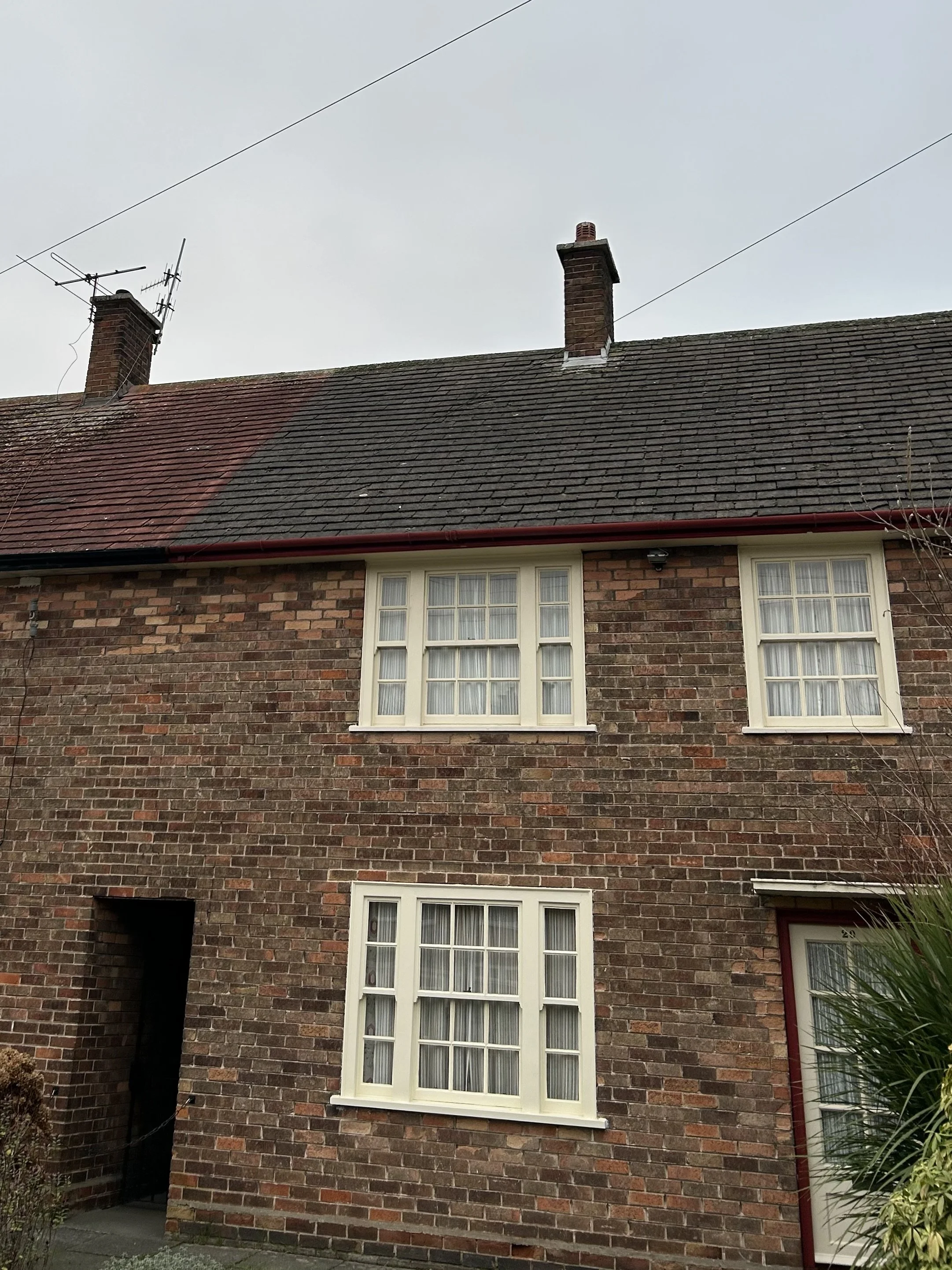 A brick house with three windows on the front, a black entrance door on the left, and a window on the right edge. The roof has two brick chimneys, and the sky is overcast.