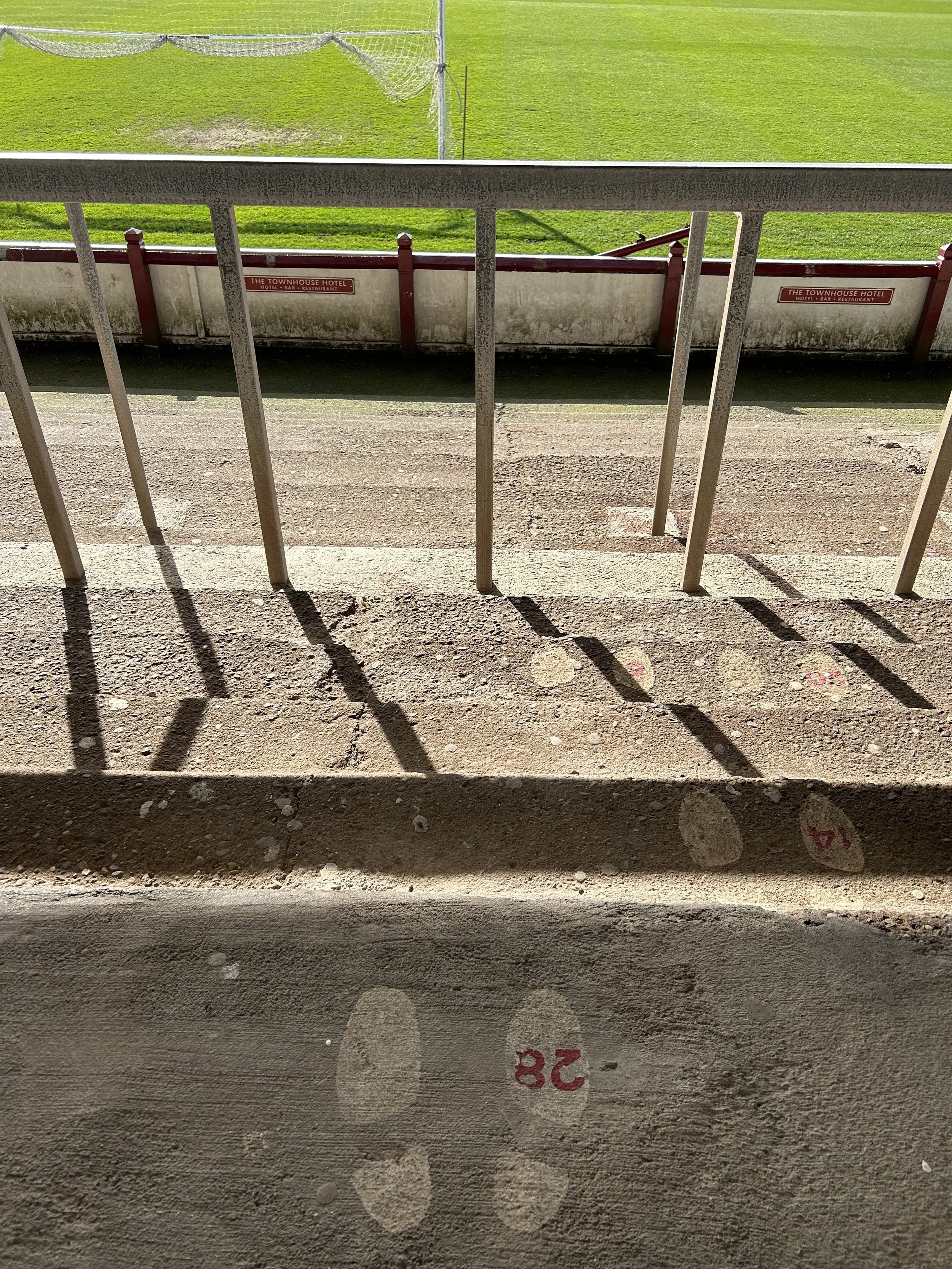View from a balcony overlooking a grassy sports field with a gravel or dirt running track. Shadows of the railing are cast on the ground. There are red and white signs on the fence that read 'The Townhouse Hotel'.