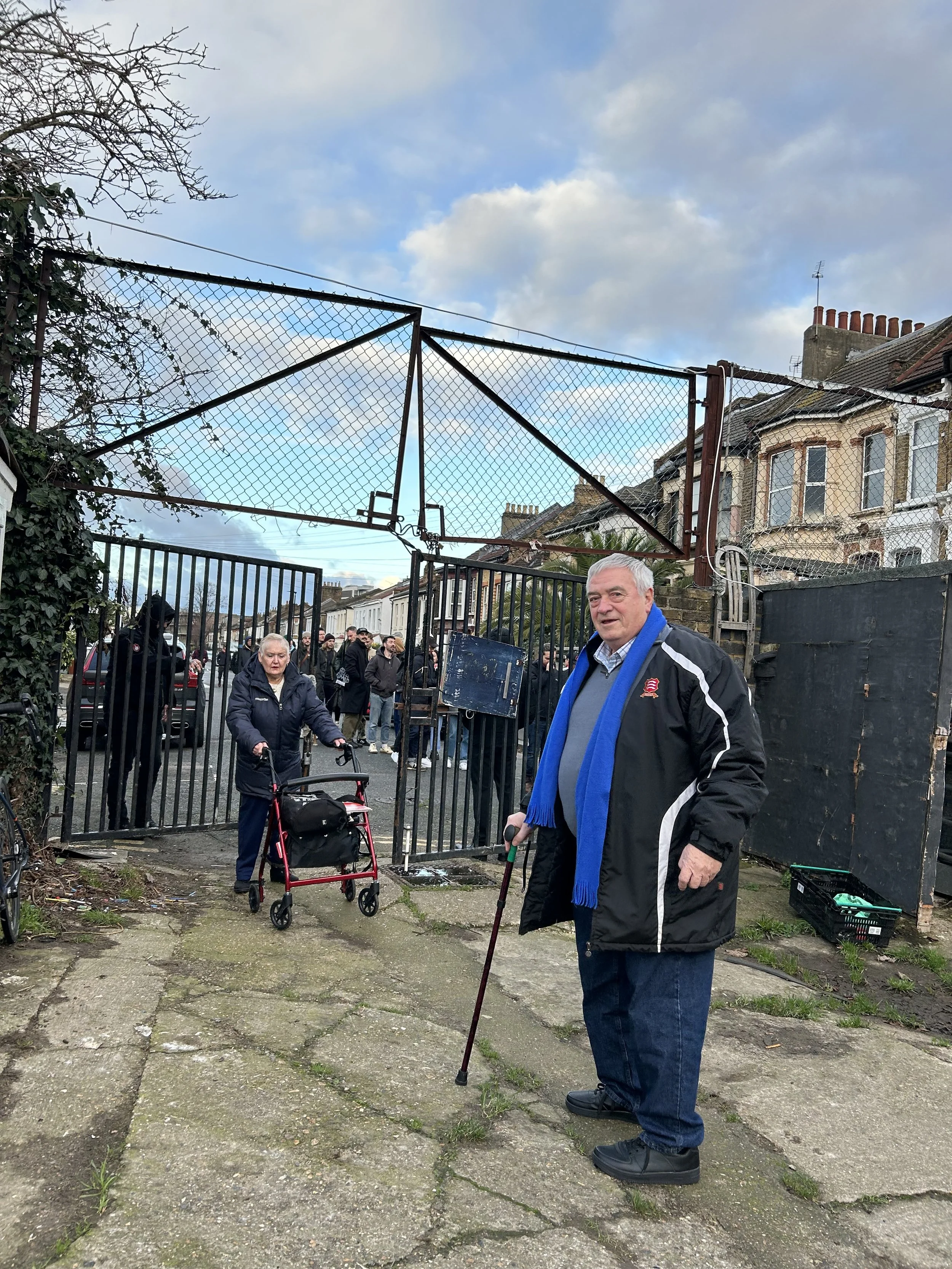 A man with gray hair standing outside, holding a cane, wearing a black jacket with a blue scarf, in front of an open gate. Behind him, a woman using a walker and a group of people are visible, with residential buildings in the background and a cloudy