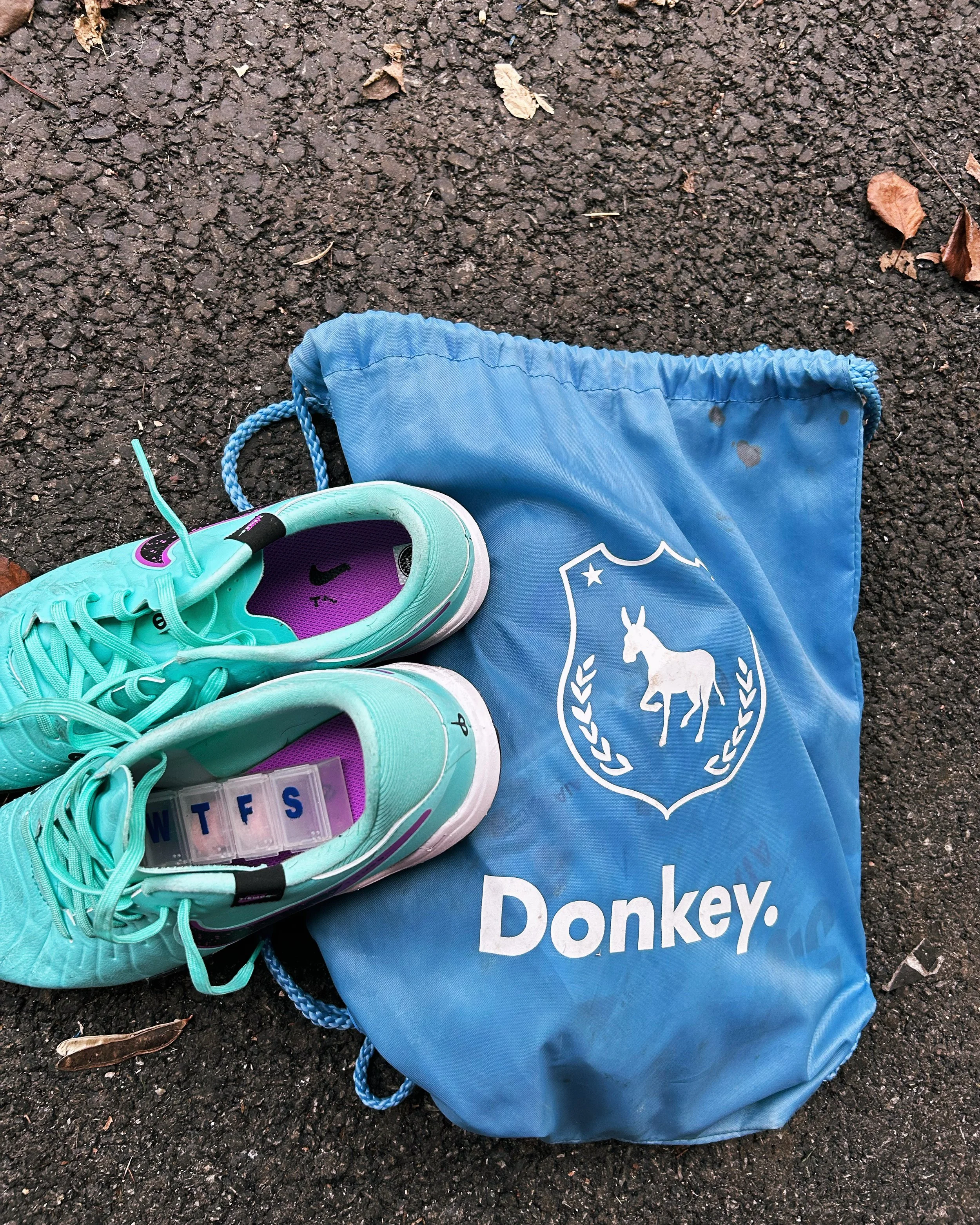 A pair of turquoise athletic shoes placed on a blue drawstring bag with a white donkey logo. The shoes have a purple insole with black writing and a transparent box with the days of the week initials inside. The shoes are on asphalt with fallen leave