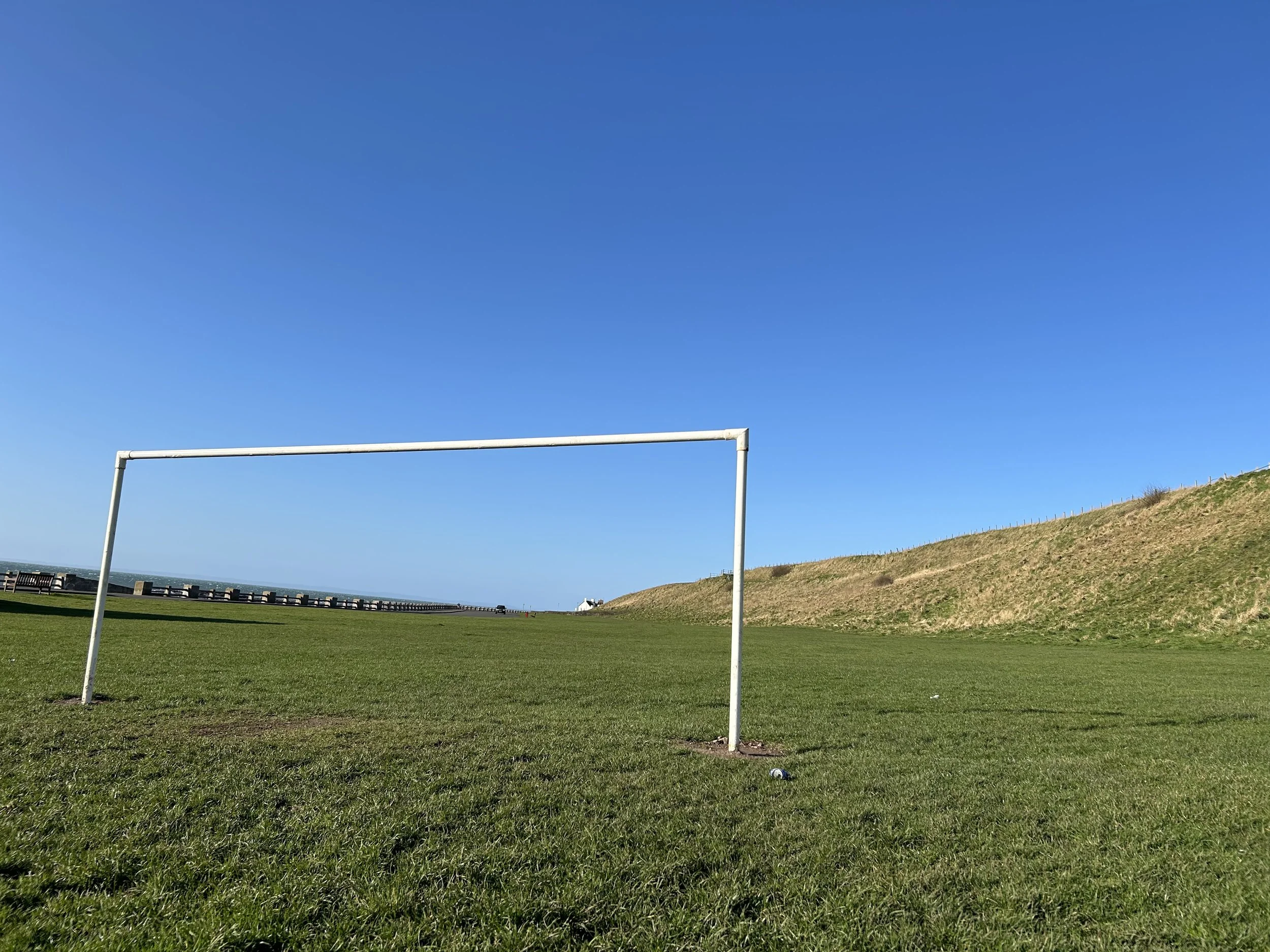 Empty soccer goal on a grassy field with a hill in the background under a clear blue sky.