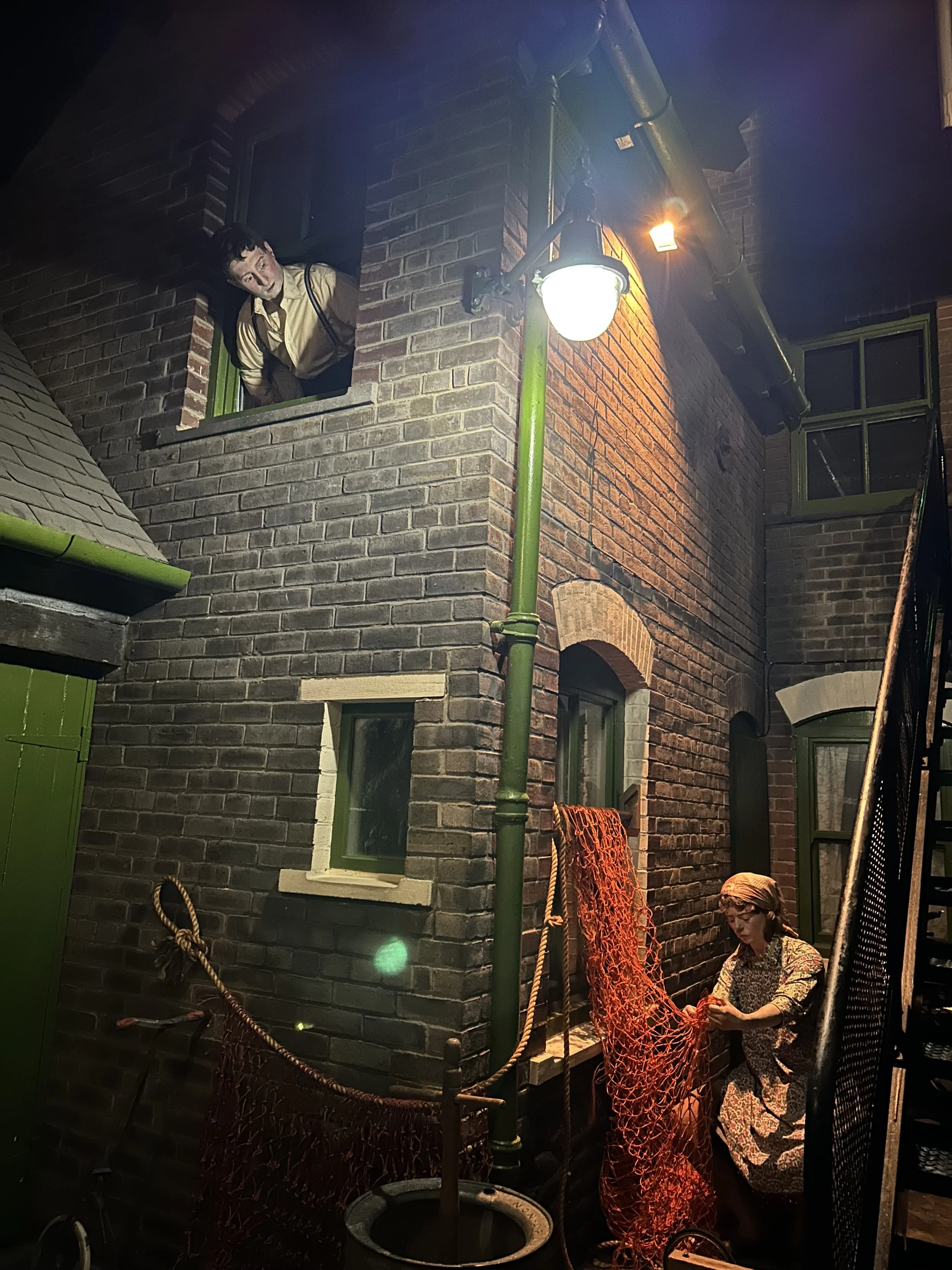 Nighttime scene of two mannequins on a street corner, one popping out of a second-story window and the other wearing a bonnet and working with an orange fishing net on the sidewalk, with brick buildings and street lamps.