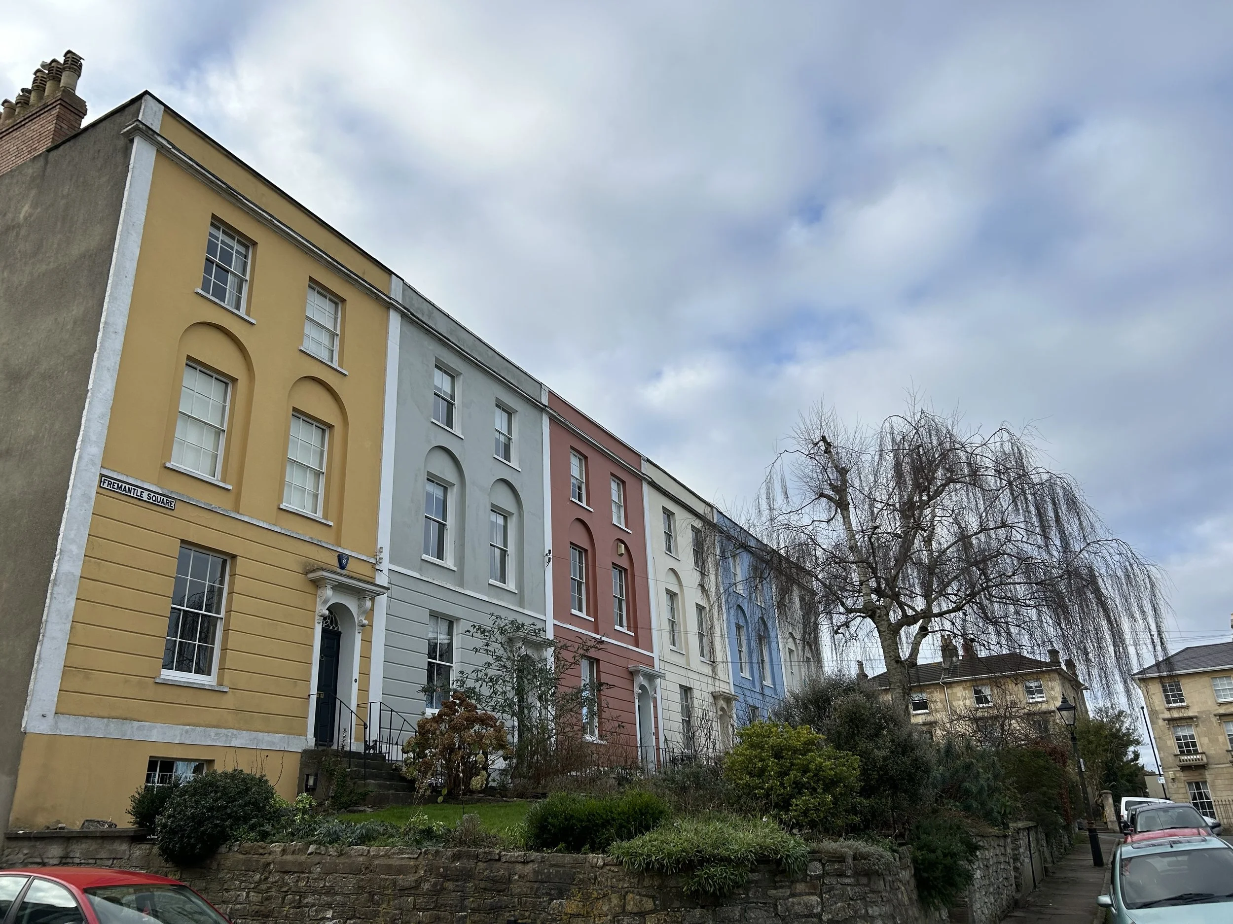 Colorful row houses on a hill with trees and parked cars, under a partly cloudy sky.