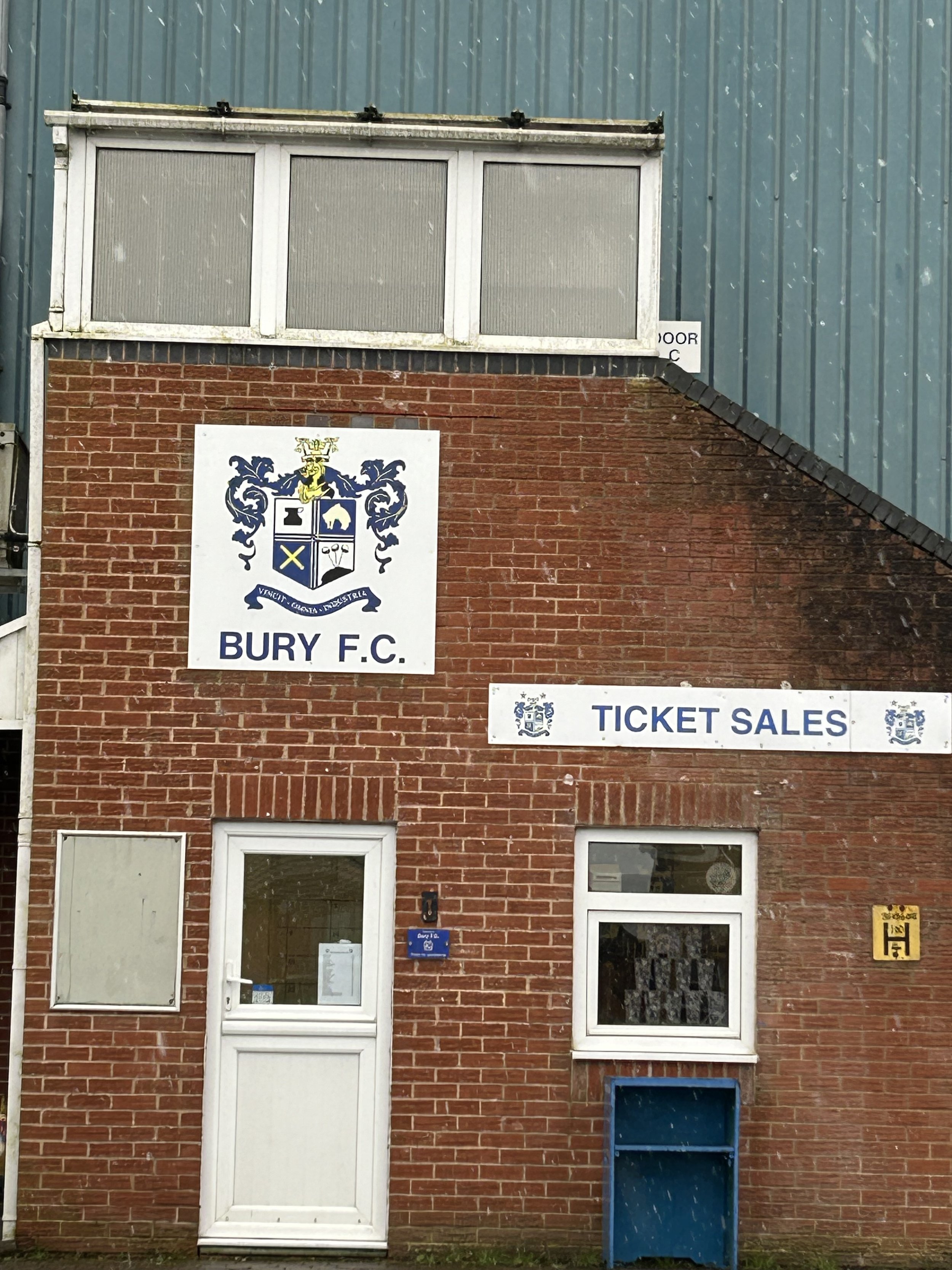 The exterior of a brick building with signs for Bury F.C. and ticket sales. The building has a white door, a window, and a blue property box, with a corner of the building visible and a clear sky behind.