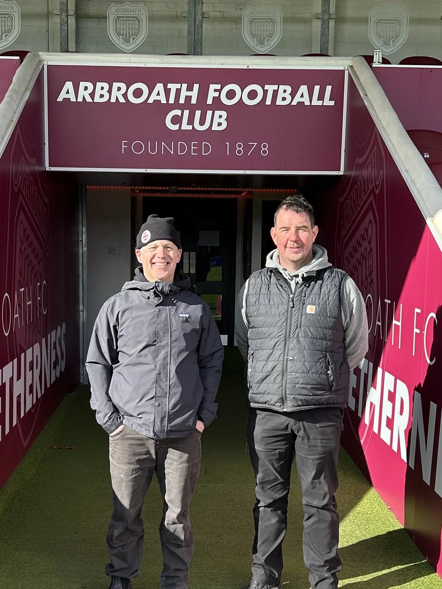 Two men standing on a green turf field outside the entrance to Arbroath Football Club, which was founded in 1878. The sign above their heads reads "ARBROATH FOOTBALL CLUB." The men are dressed in casual outdoor clothing, with one wearing a beanie hat