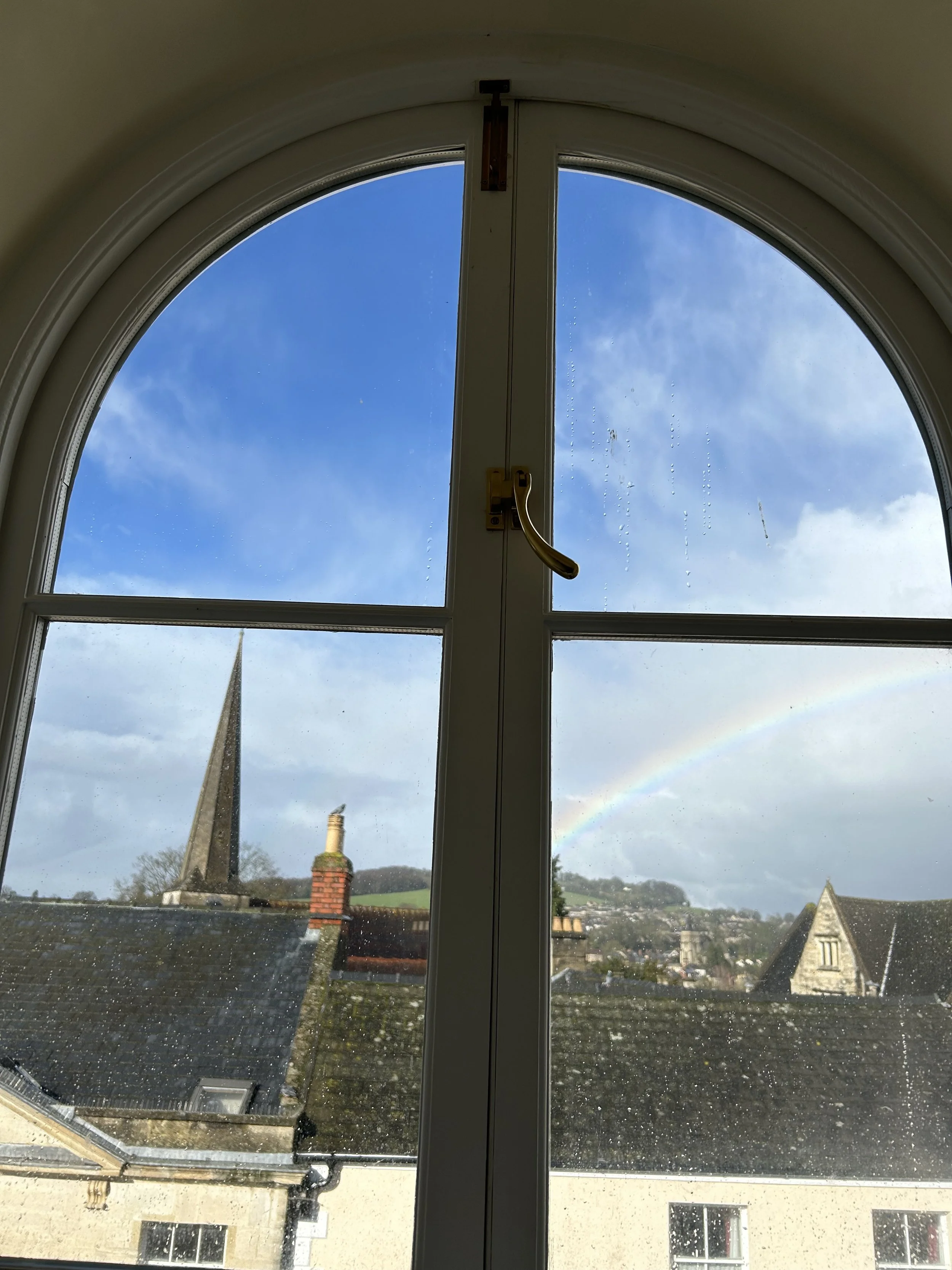 View through a window showing rooftops, a church steeple, and a rainbow in a partly cloudy sky.