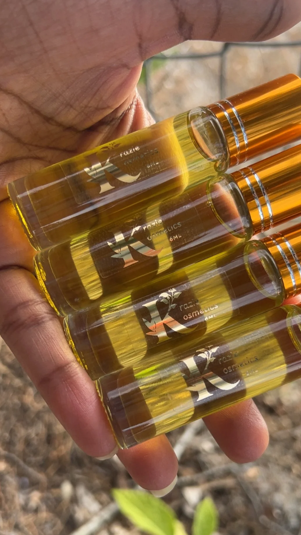Close-up of a person's hand holding four small bottles of golden yellow liquid, labeled 'RXCOSMETICS 6ML,' outdoors with a blurred natural background.