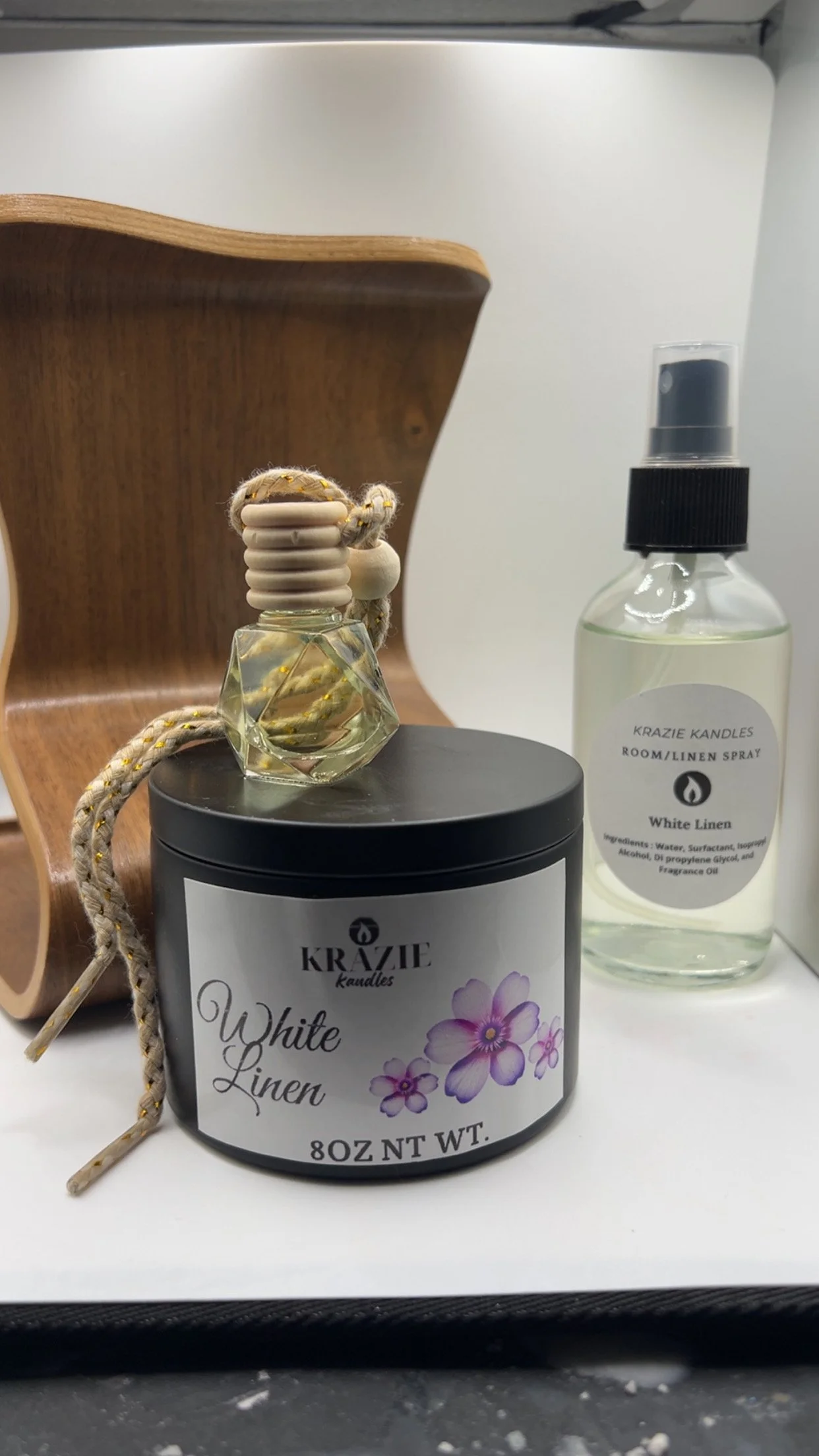 A black candle jar labeled 'KRAZIE candles White Linen' with purple flower design, a clear glass perfume bottle with a white cap and a braided rope, and a spray bottle labeled 'KRAZIE KANDLES ROOM/LINEN SPRAY White Linen' on a white surface with a wooden item behind them.