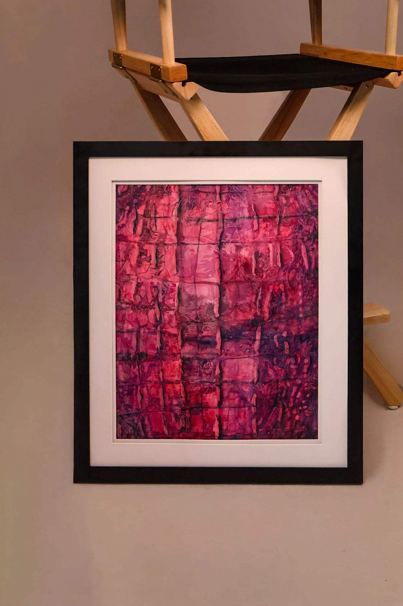 A framed abstract art piece with pink and purple tones hangs on a beige wall, with a wooden folding chair positioned above it.