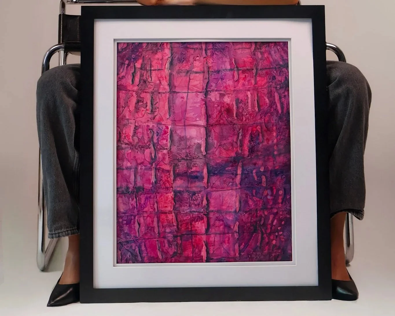 Abstract pink and purple textured artwork in a black frame, being held by a person sitting on a chair with only their legs and feet visible.