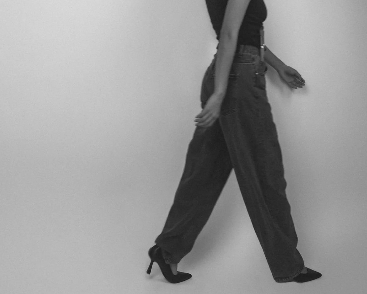 Black and white photo of a woman walking with high heels, wearing loose-fitting jeans and a sleeveless top, against a plain white background.