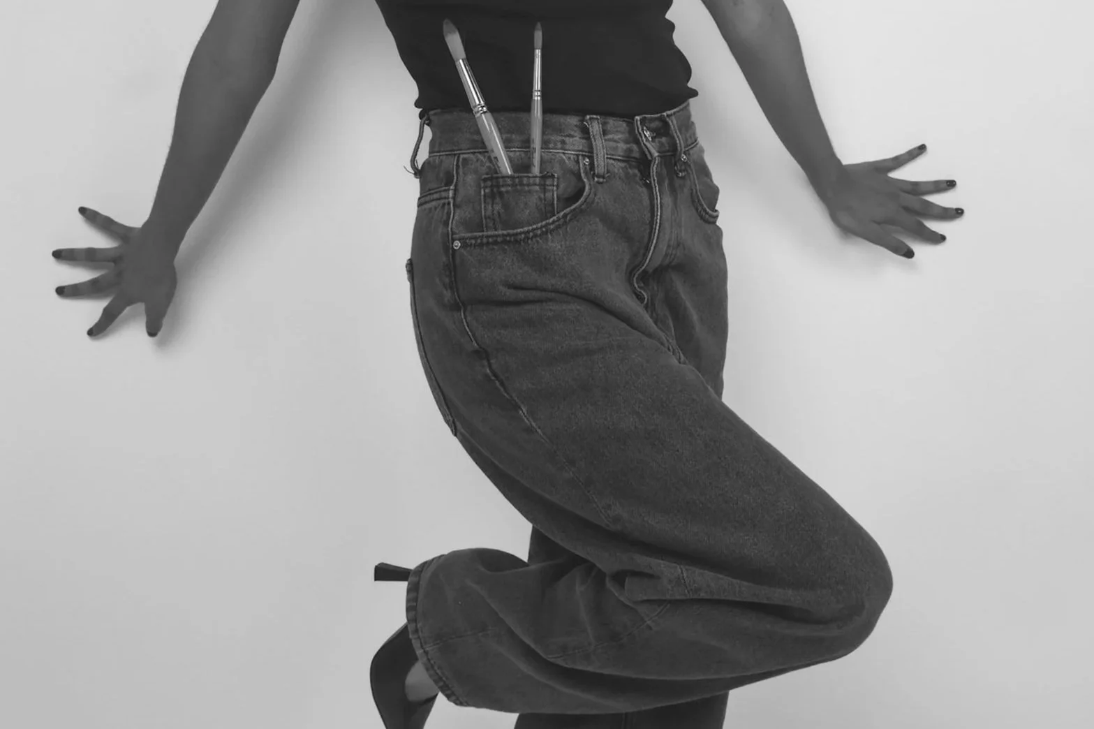 Close-up of a person in high heels and casual jeans, leaning against a plain wall, with two brushes tucked into their waistband.
