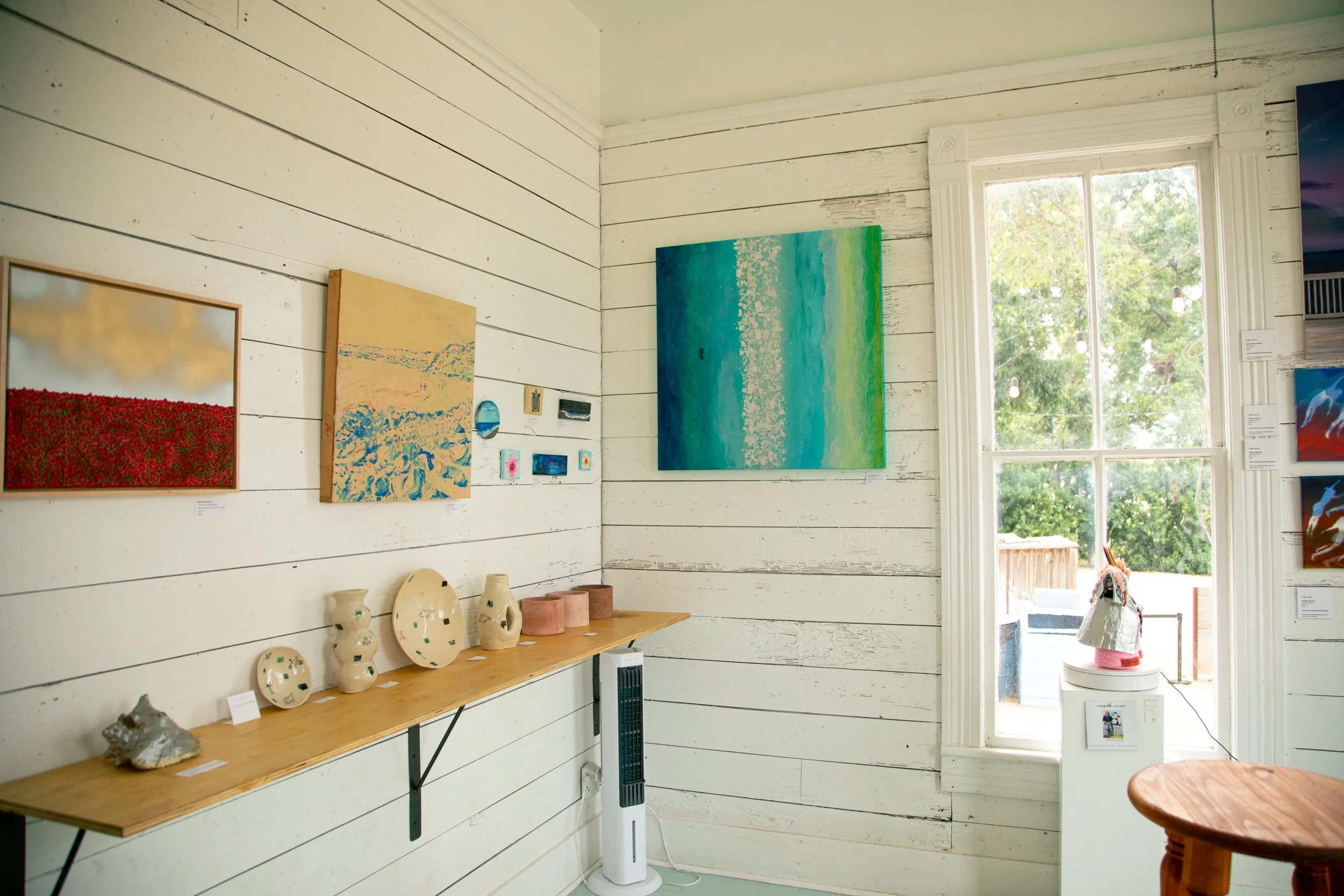 Interior of an art gallery with white wooden walls, showcasing colorful paintings and ceramic pottery on a wooden shelf, bright natural light from a window on the right.