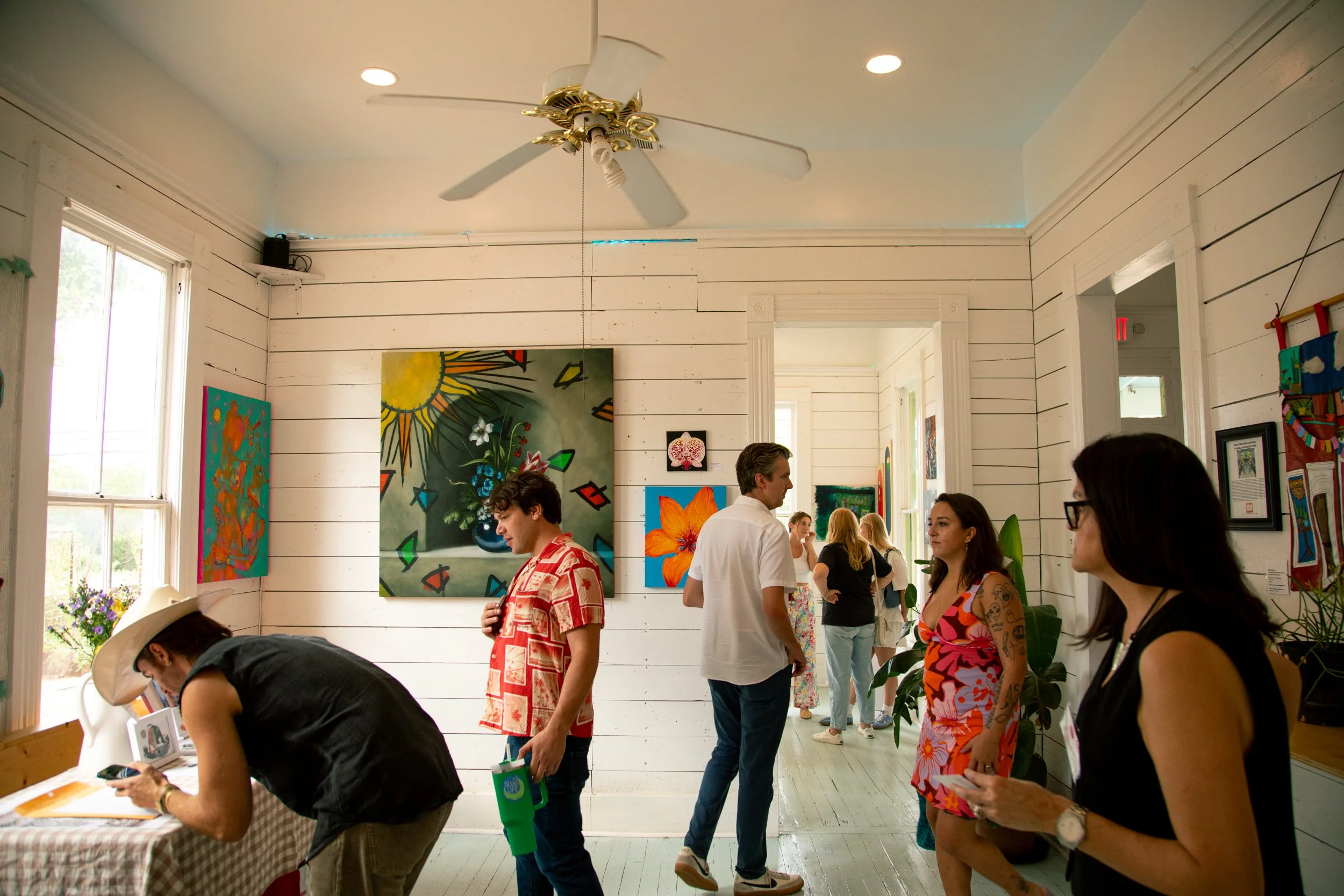 People viewing artwork in an art gallery with white paneled walls, colorful paintings, and a ceiling fan.