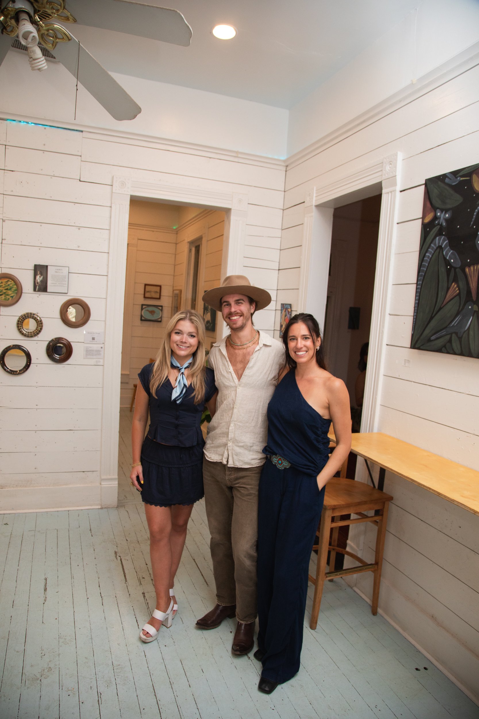 A group of three people posing indoors in front of a white wall with wall art, smiling at the camera.