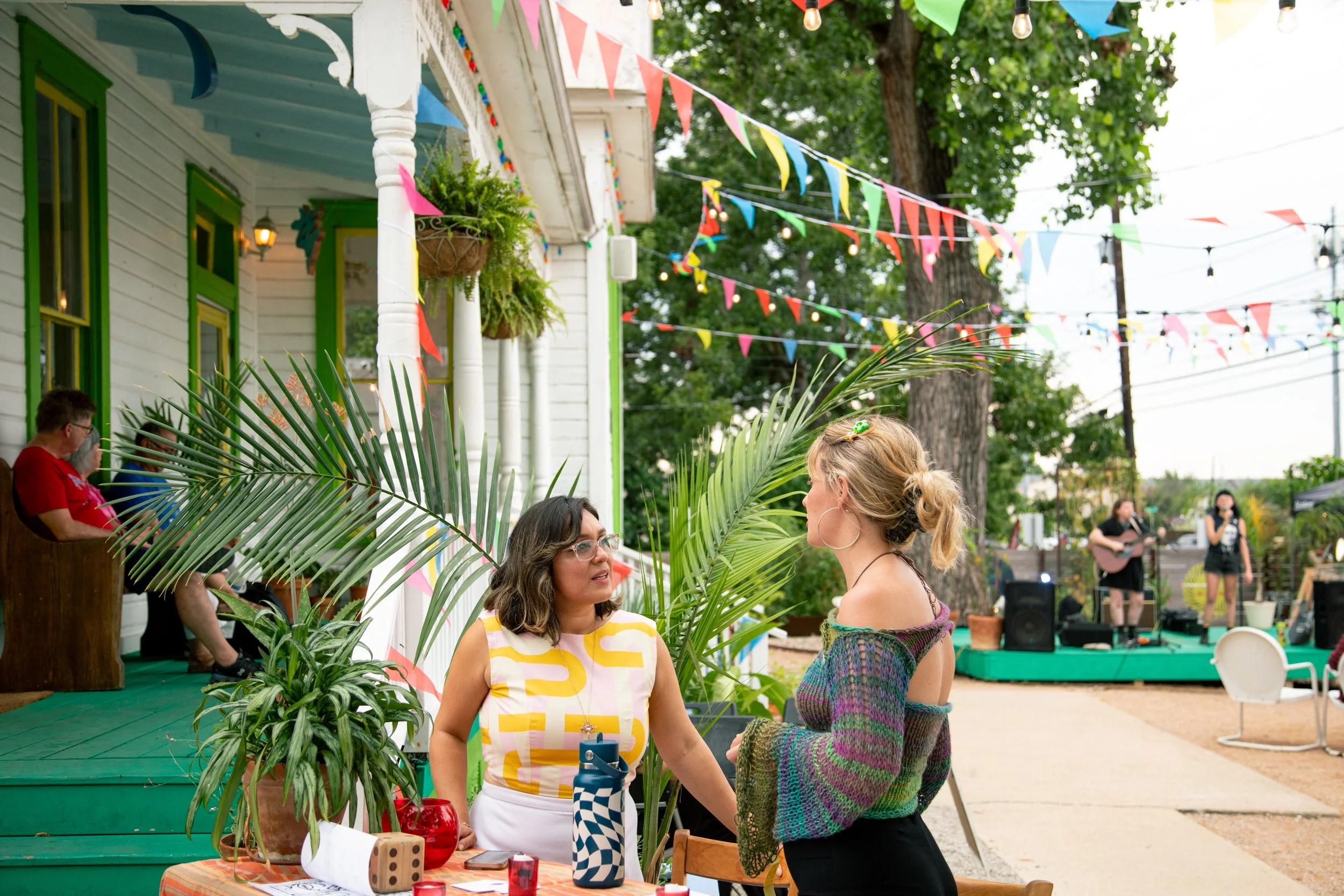 Two women are engaged in conversation at an outdoor celebration with string lights and colorful bunting. One woman is sitting at a table with potted plants, and the other is standing. In the background, a band is performing on a small stage.