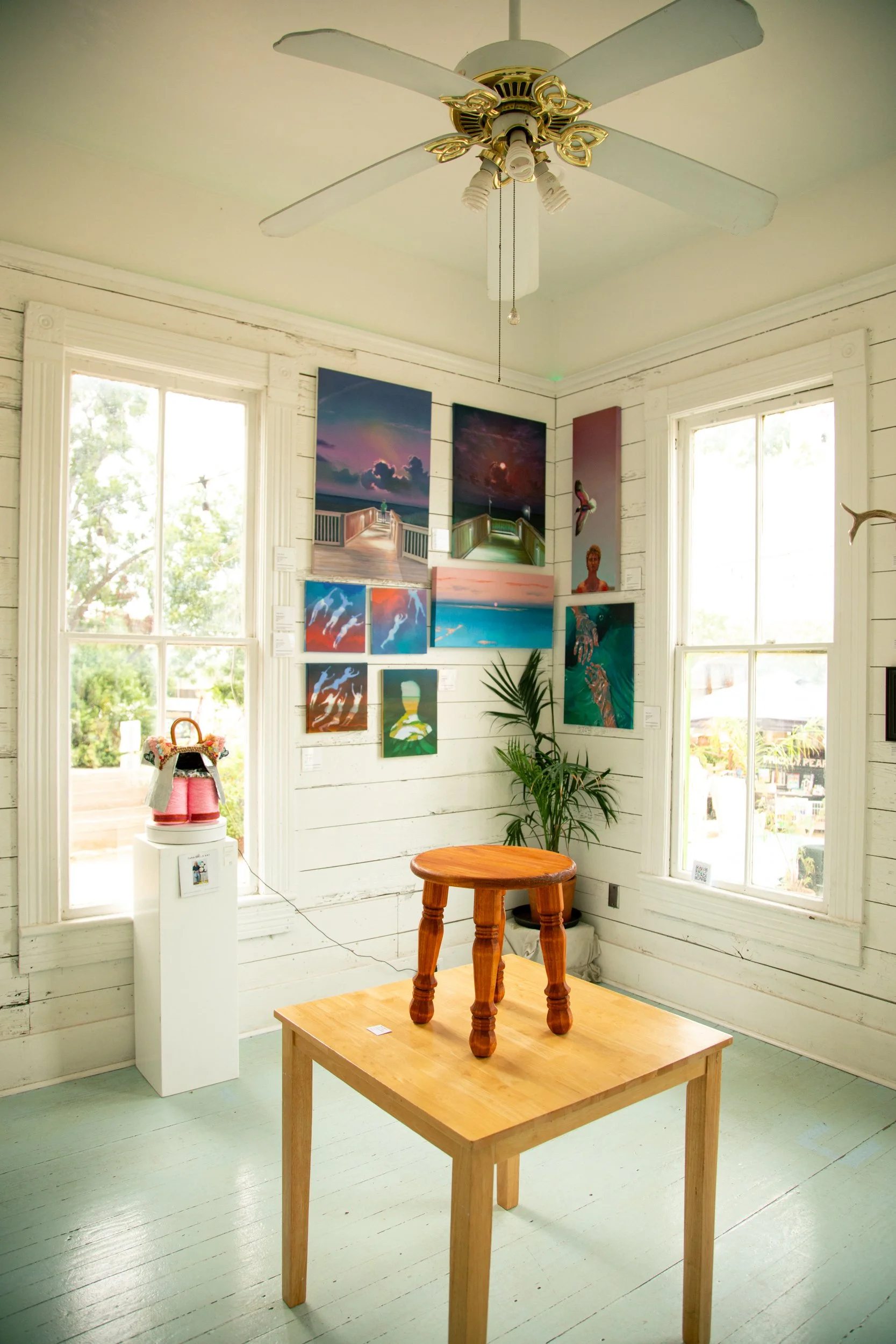 An art gallery room with white wooden walls and floors, featuring a collection of colorful paintings on the wall, a small wooden table with a small wooden sculpture on top, and a ceiling fan with four blades.
