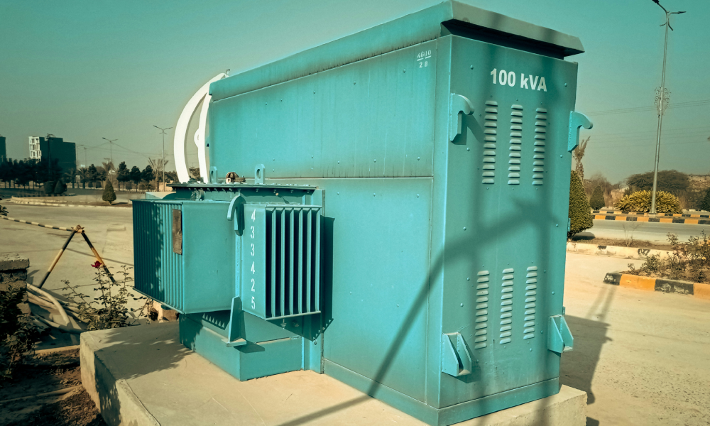 A large electrical transformer with a label indicating 100 kVA capacity, situated outdoors on a concrete base with a blue-green color.