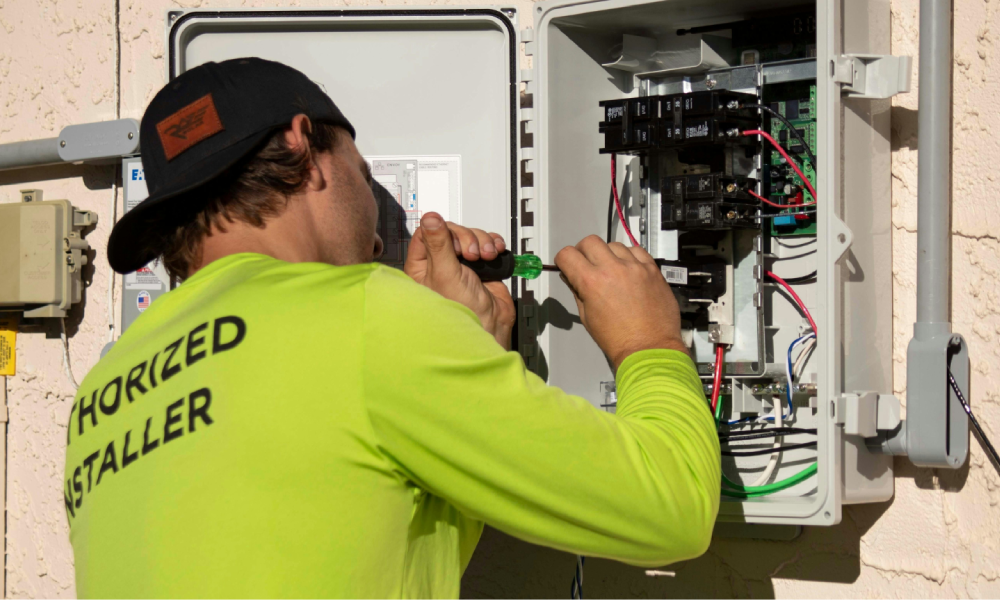 Technician working on an electrical panel outdoors, wearing a bright yellow shirt with 'HORIZONED INSTALLER' written on the back, using a screwdriver.