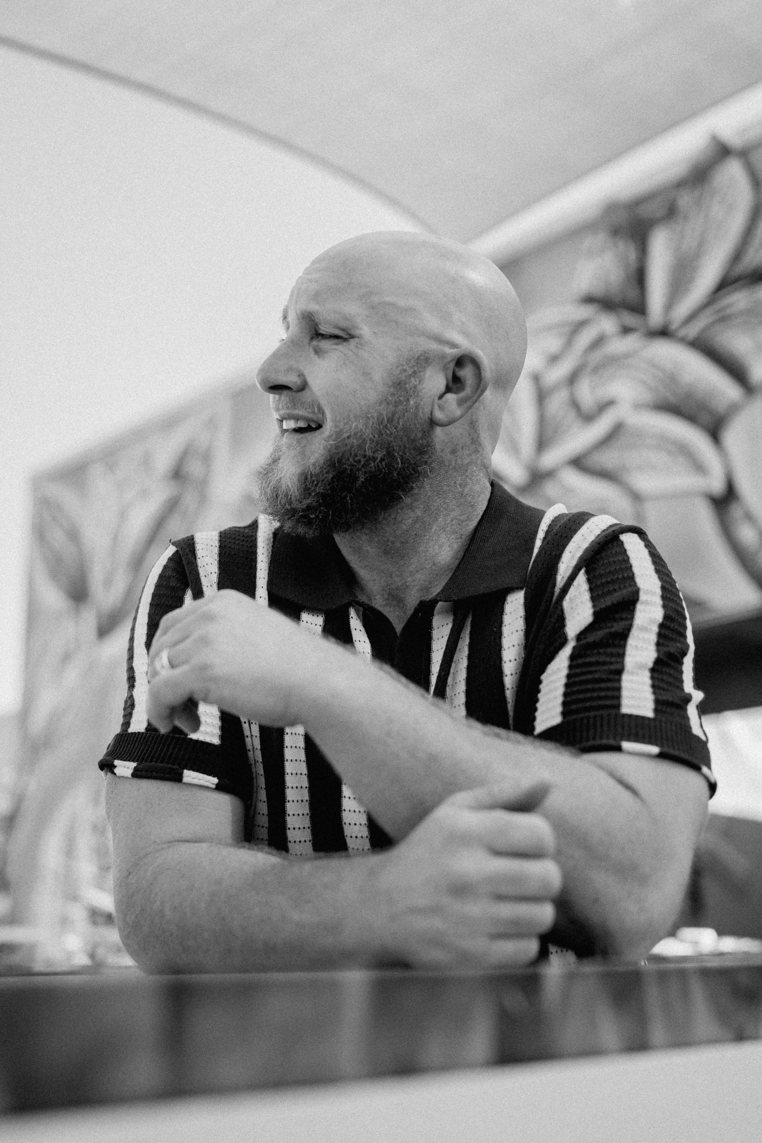 Black-and-white photo of a smiling man with a beard and shaved head, wearing a striped shirt, resting his arms on a table.