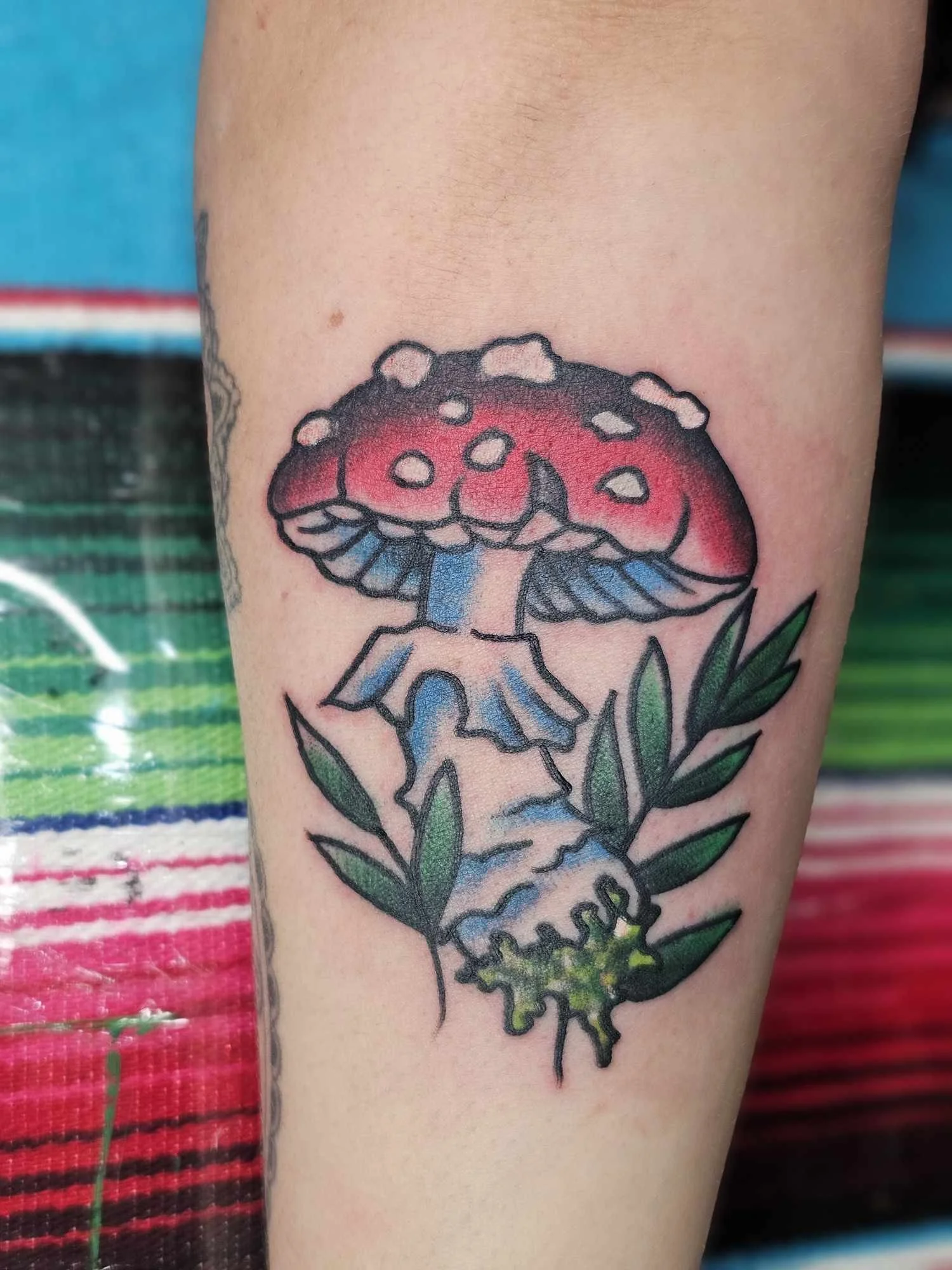 Colorful tattoo of a mushroom with a red cap and white spots, surrounded by green leaves on skin.