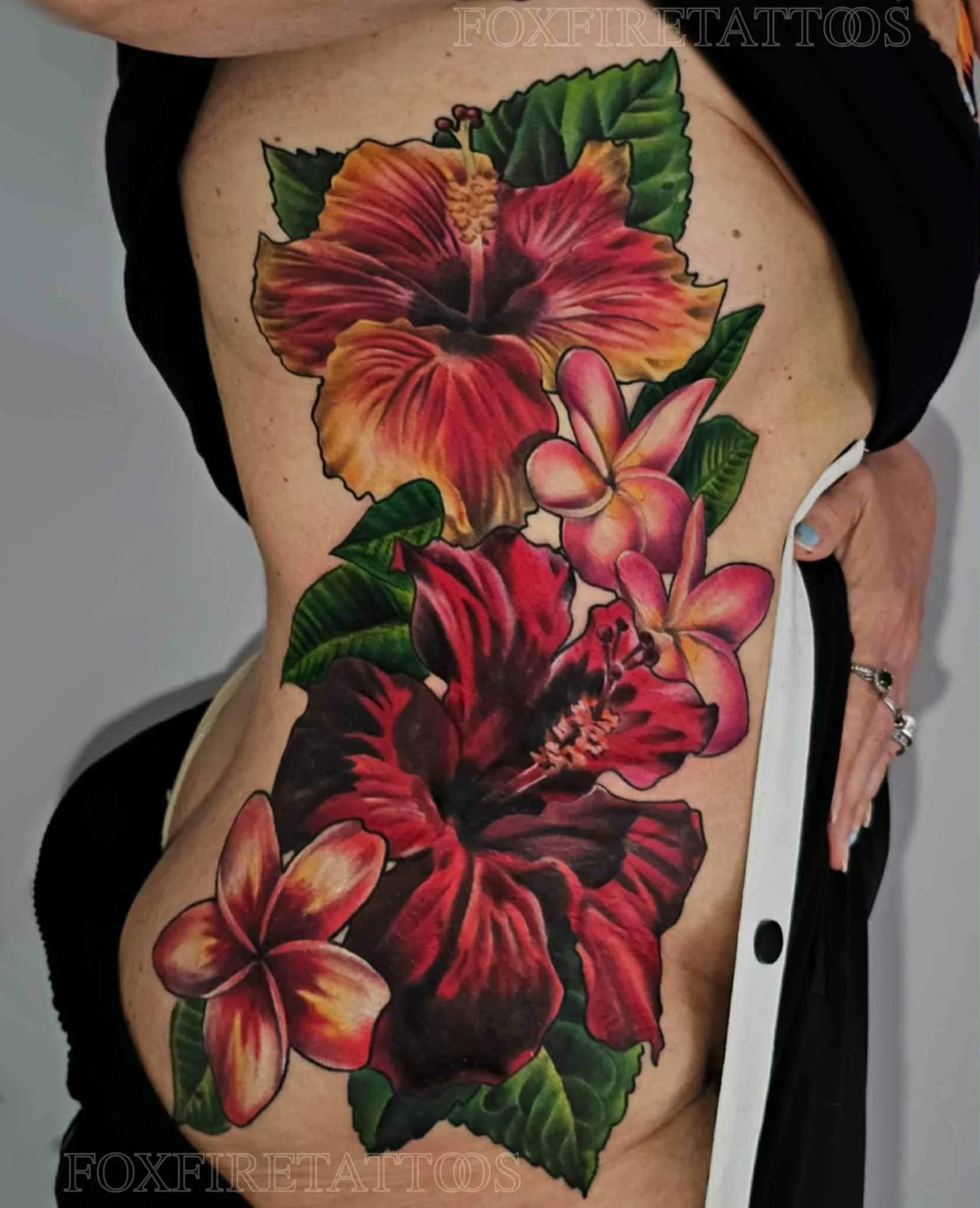Colorful floral tattoo featuring hibiscus and plumeria flowers with green leaves on a person's side torso.