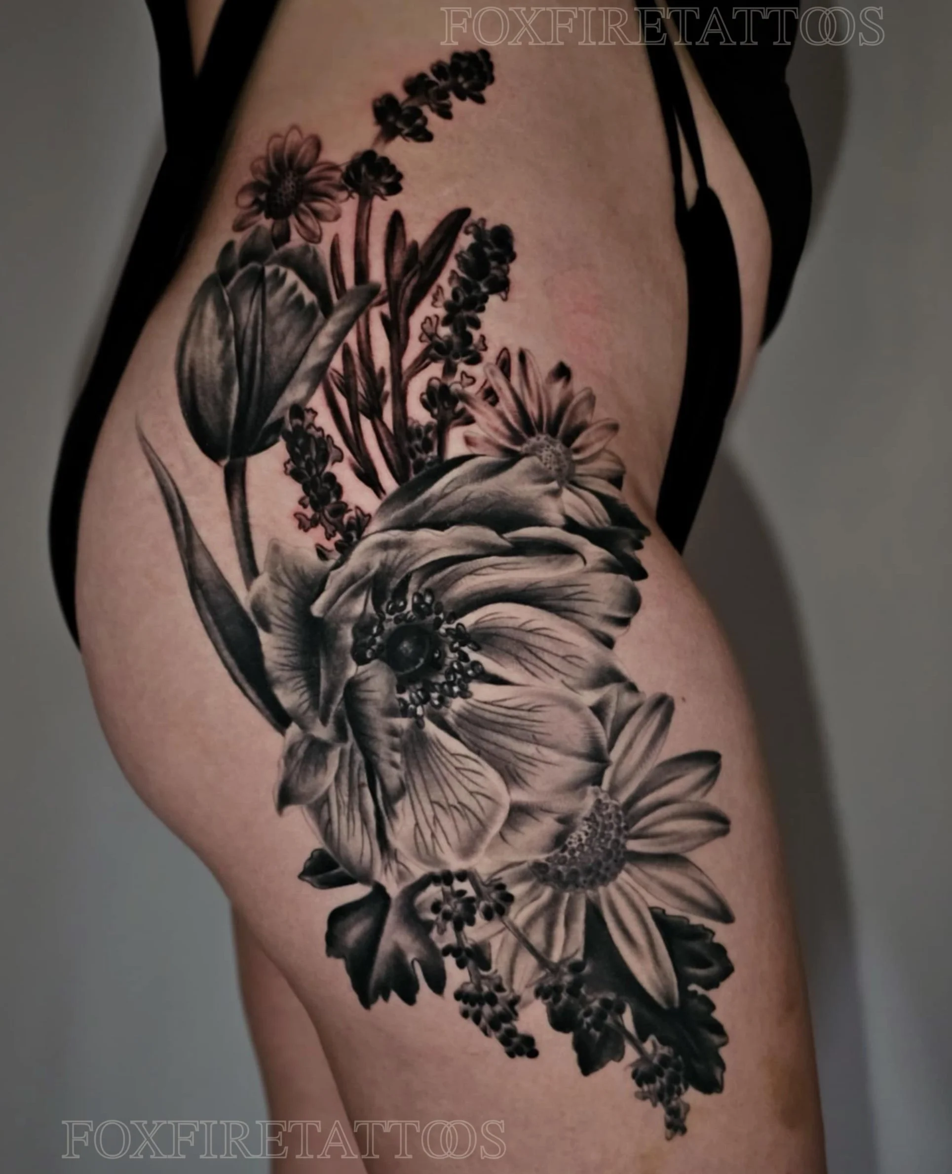 Black and gray floral tattoo on upper thigh featuring large roses, daisies, tulips, and smaller flowers with detailed shading.