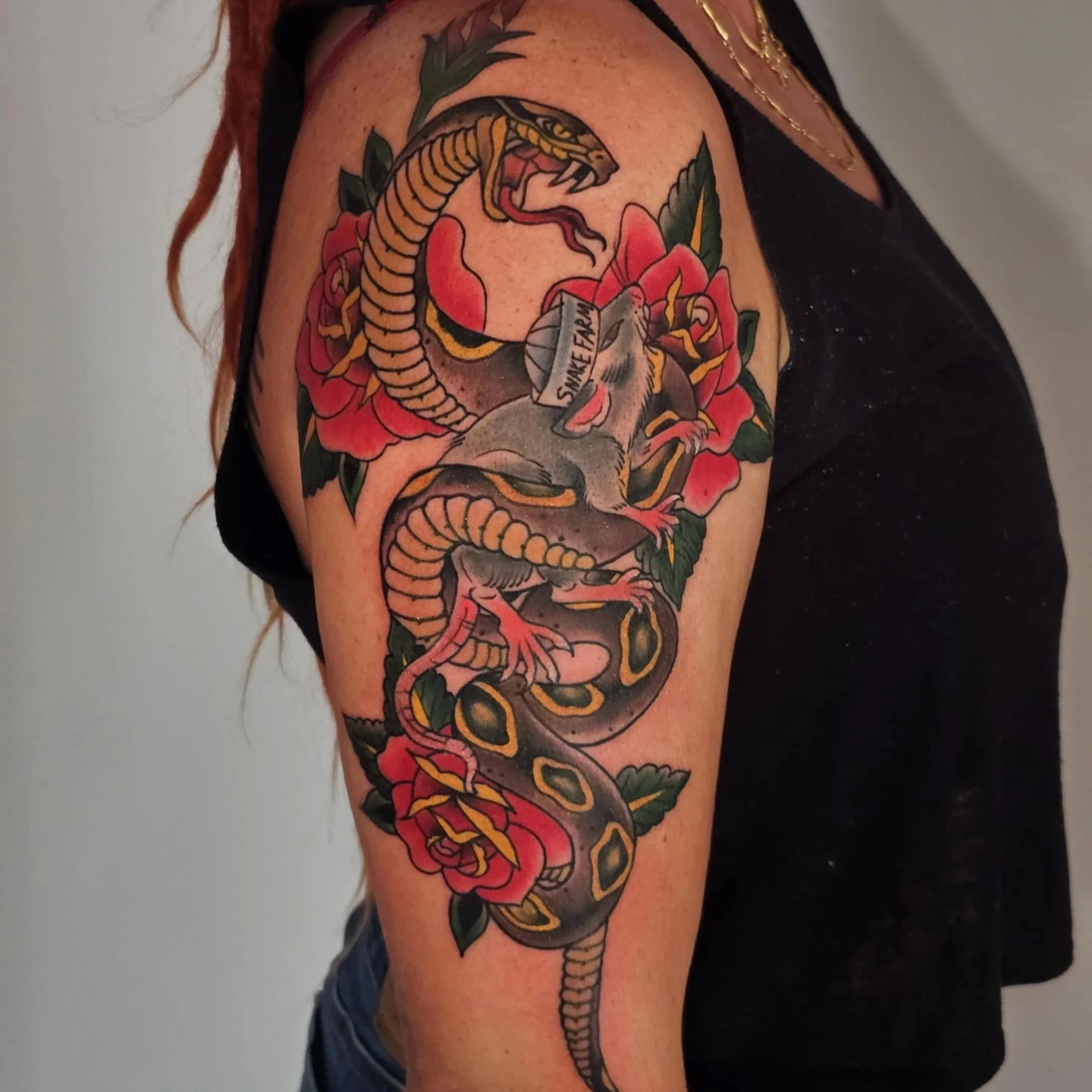 Tattoo on upper arm featuring a snake, a mouse with a syringe labeled "Snake Fret," and red roses with green leaves.