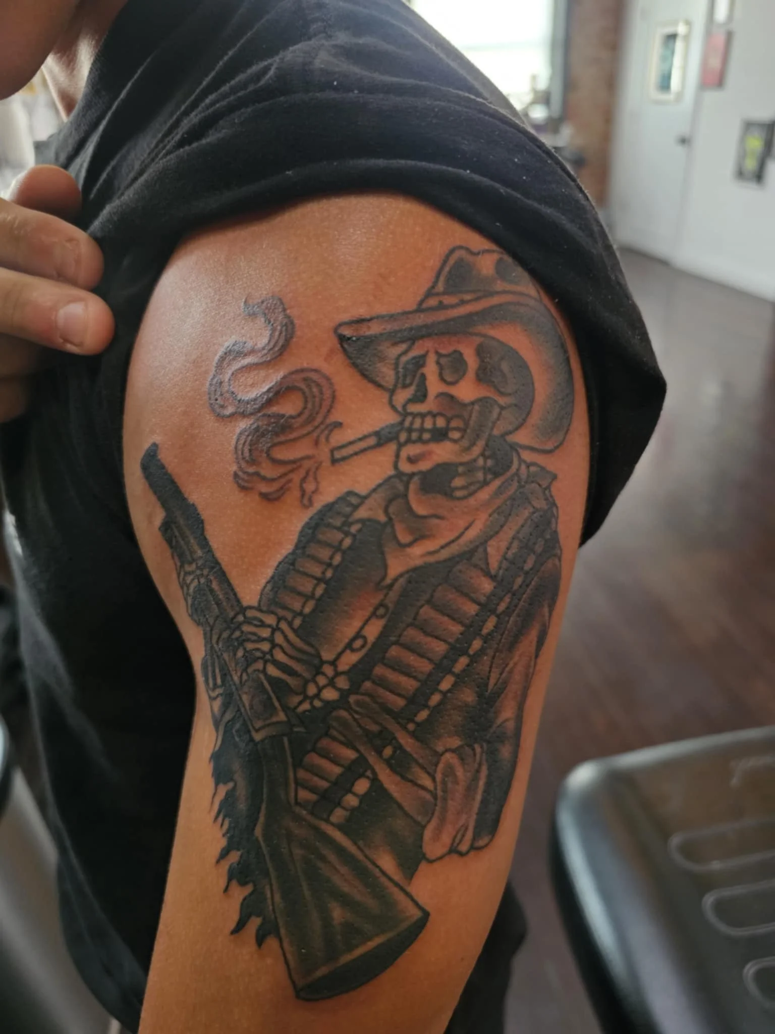 Tattoo of a skeleton cowboy wearing a wide-brimmed hat and bandana, smoking a cigar, with a large firearm across his chest.