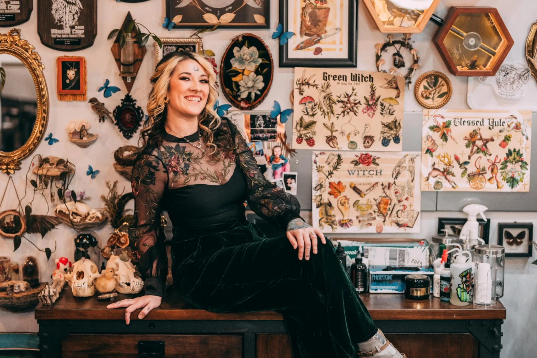 A woman with blonde hair, tattoos, and a moon tattoo on her forehead, smiling while sitting on a wooden table in a room decorated with tattoo artwork, butterflies, skulls, and eclectic art.