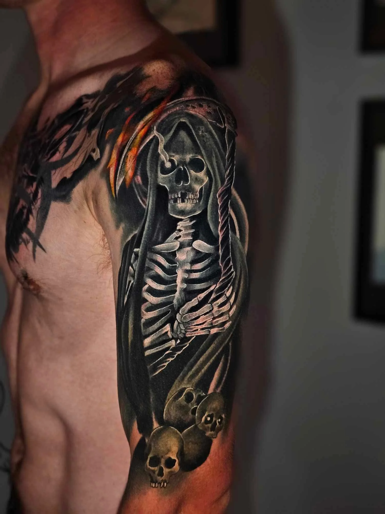 Tattoo of a skeleton with a skull mask and a scythe on an arm, with small skulls at the bottom.